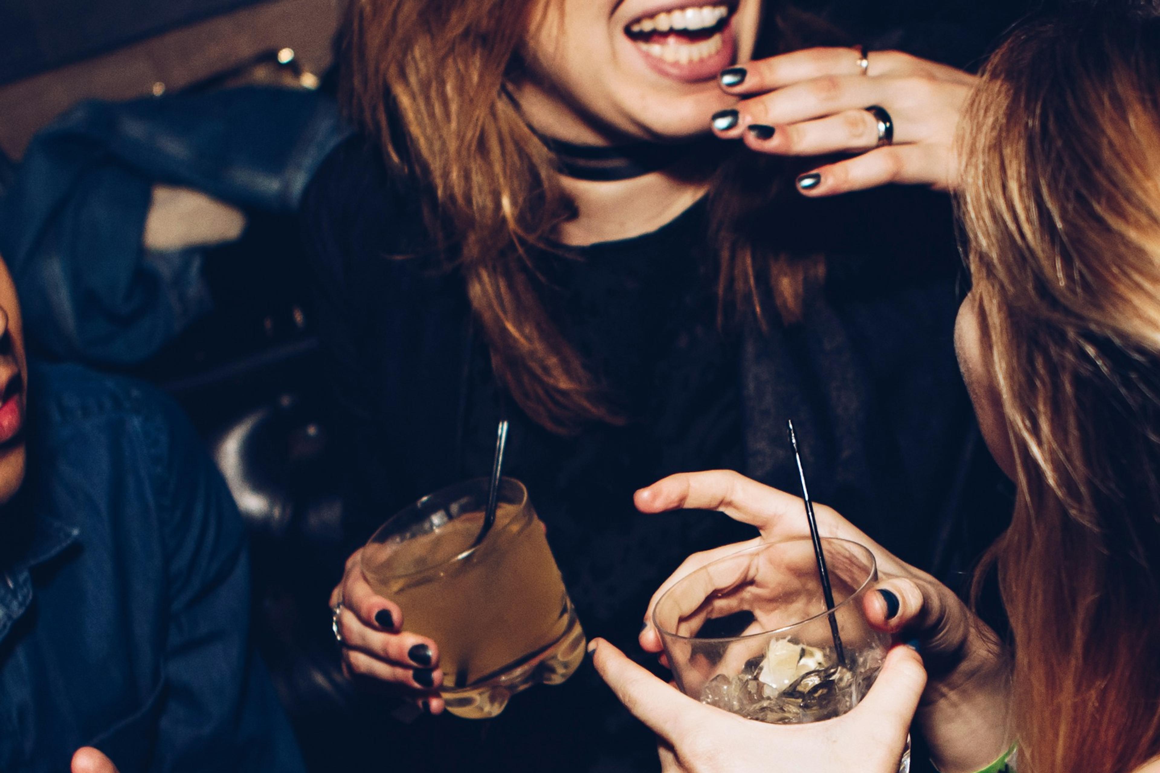 Three people laughing and holding drinks at a party, showing hands and faces partially, with a joyful atmosphere.