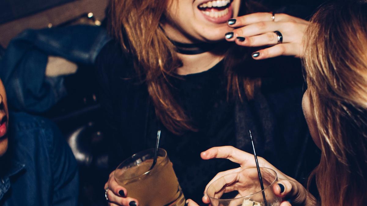 Three people laughing and holding drinks at a party, showing hands and faces partially, with a joyful atmosphere.
