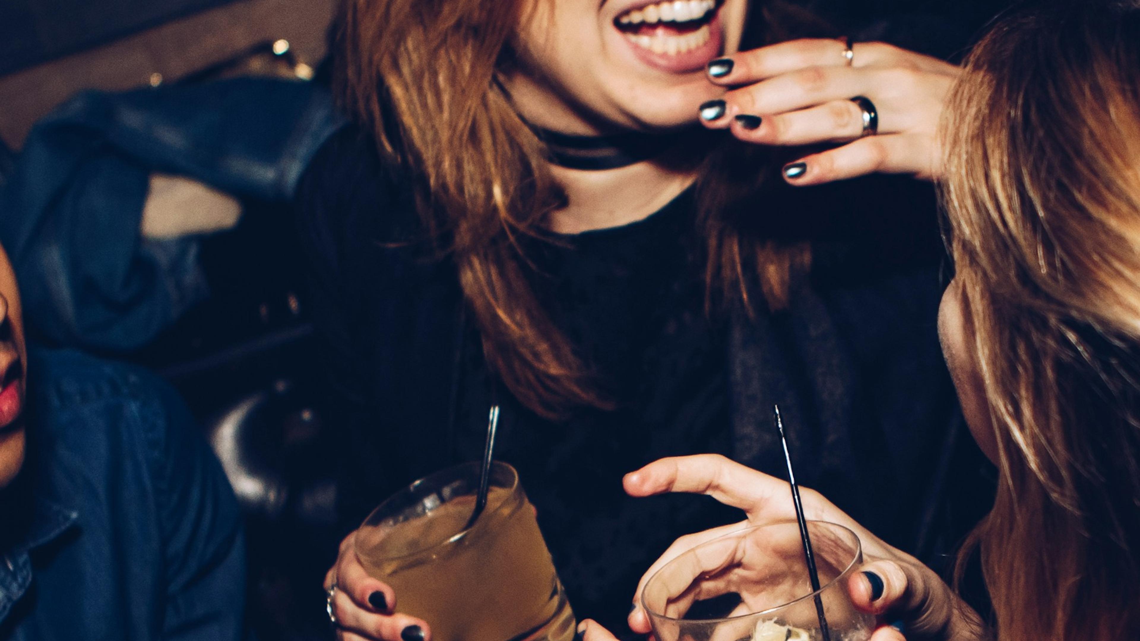 Three people laughing and holding drinks at a party, showing hands and faces partially, with a joyful atmosphere.