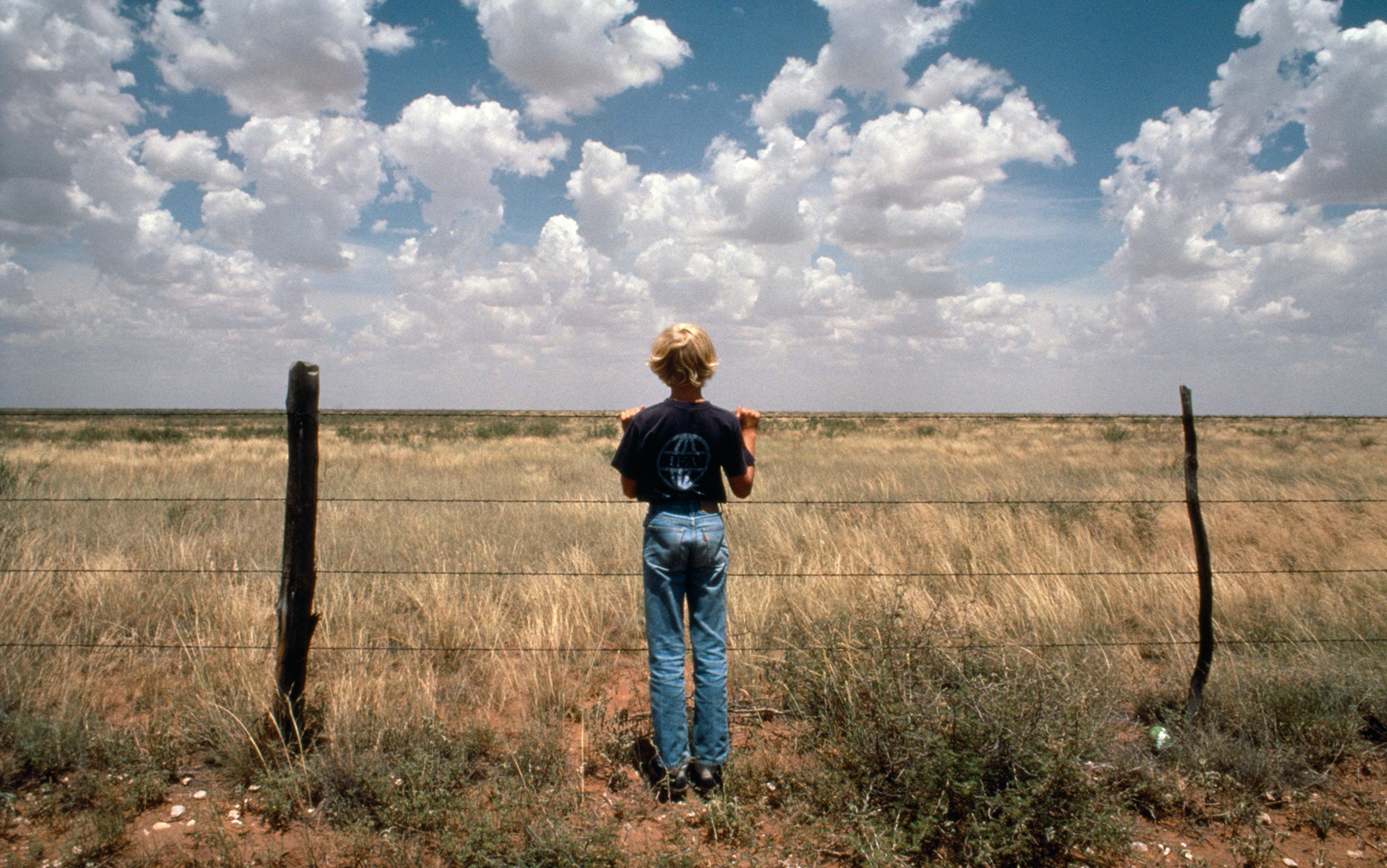 A child standing near a wire fence in an open grassy field under a blue sky with scattered clouds, viewed from behind.