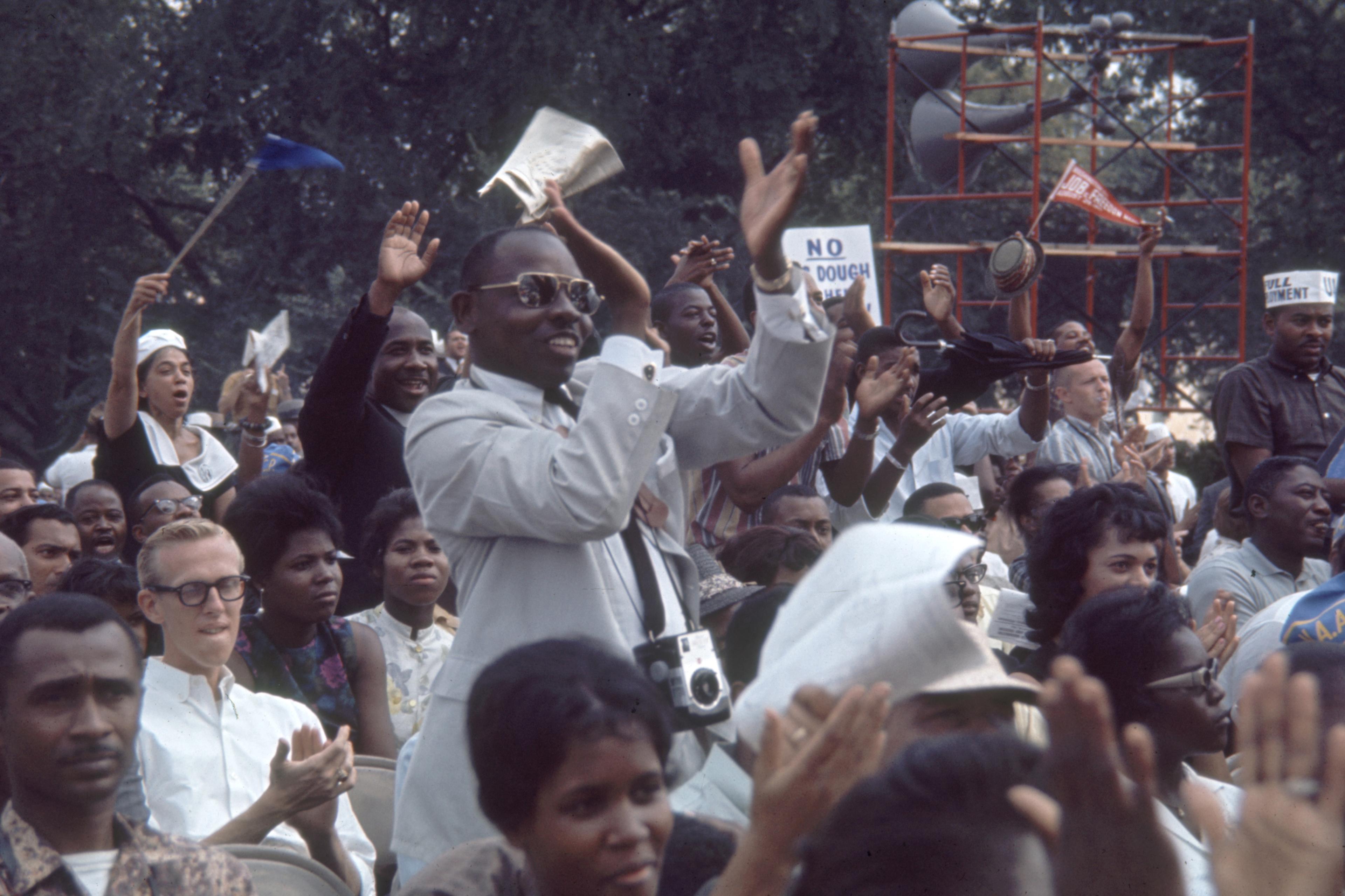 Photo of a diverse crowd clapping and cheering outdoors, with some holding signs and cameras, trees in the background.