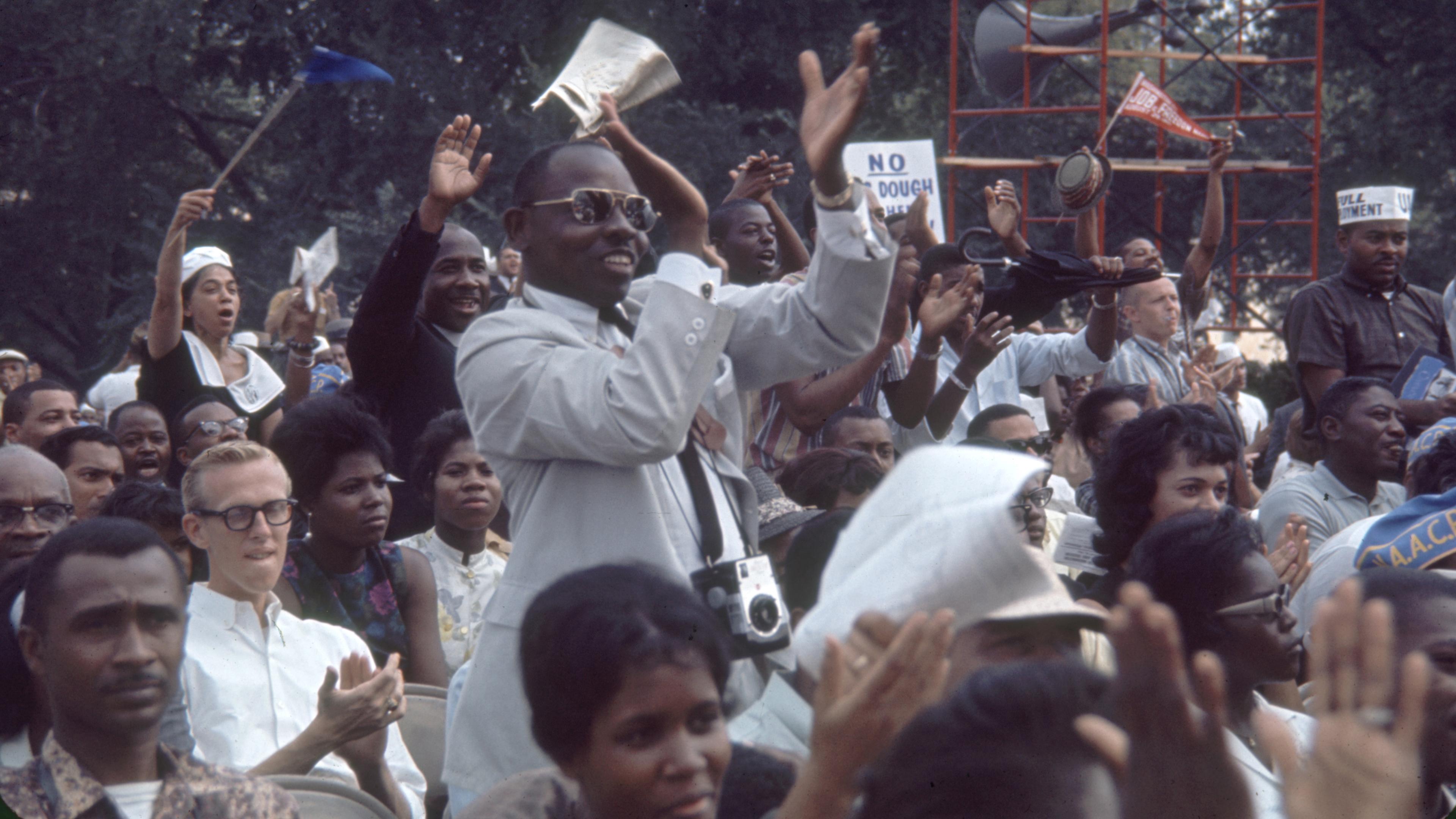 Photo of a diverse crowd clapping and cheering outdoors, with some holding signs and cameras, trees in the background.