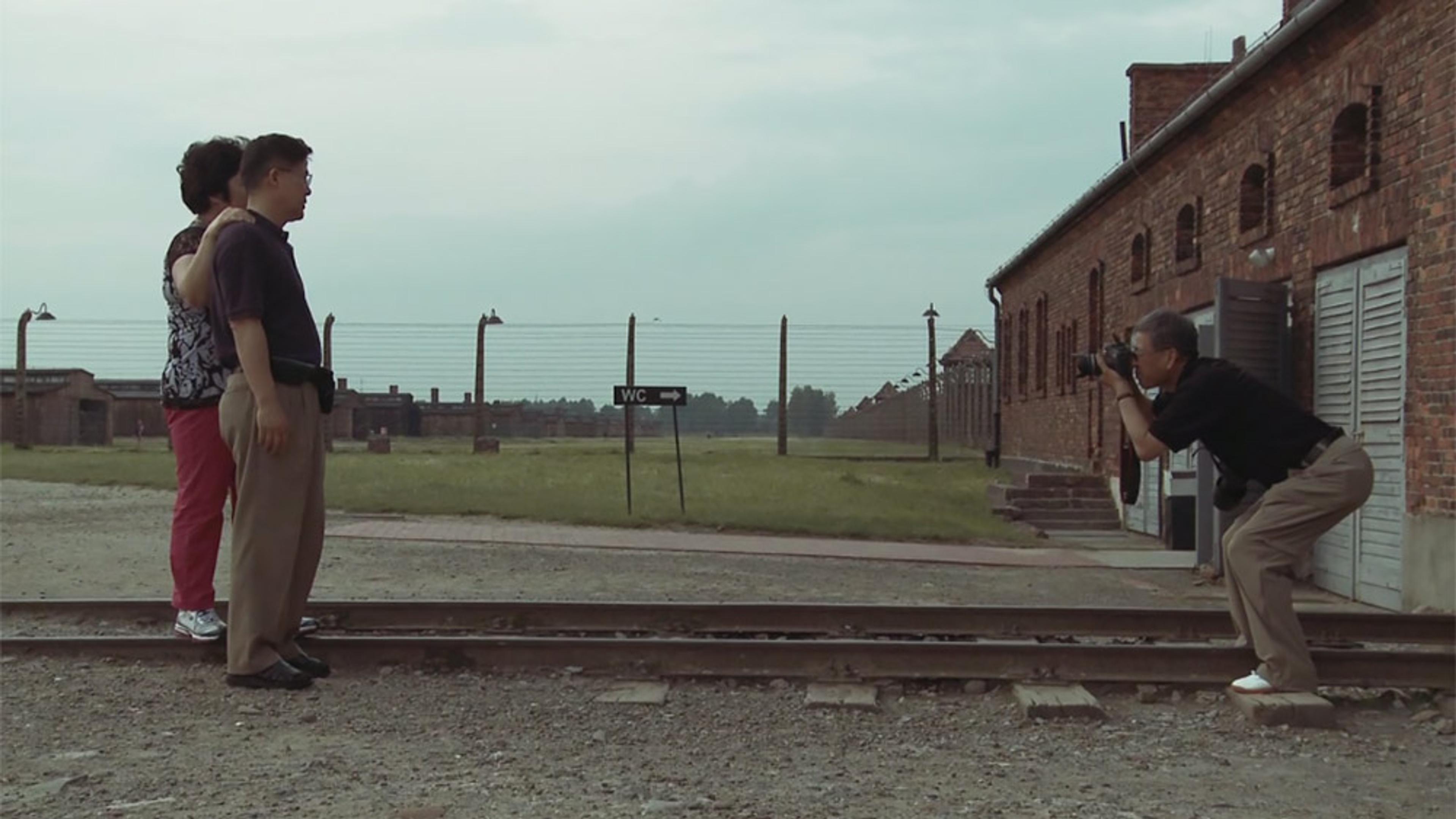 A man photographing a couple in front of a brick building and barbed wire fence at a historical or memorial site.