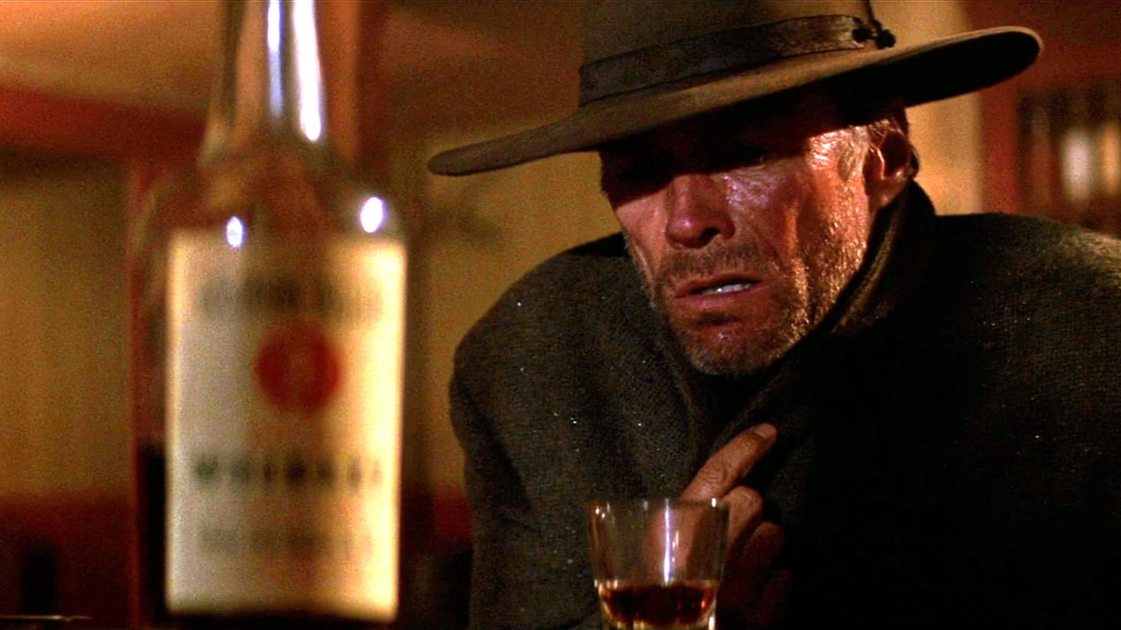 Scene from a movie with a rugged man in a hat holding his collar, a glass and bottle of whisky on the table.