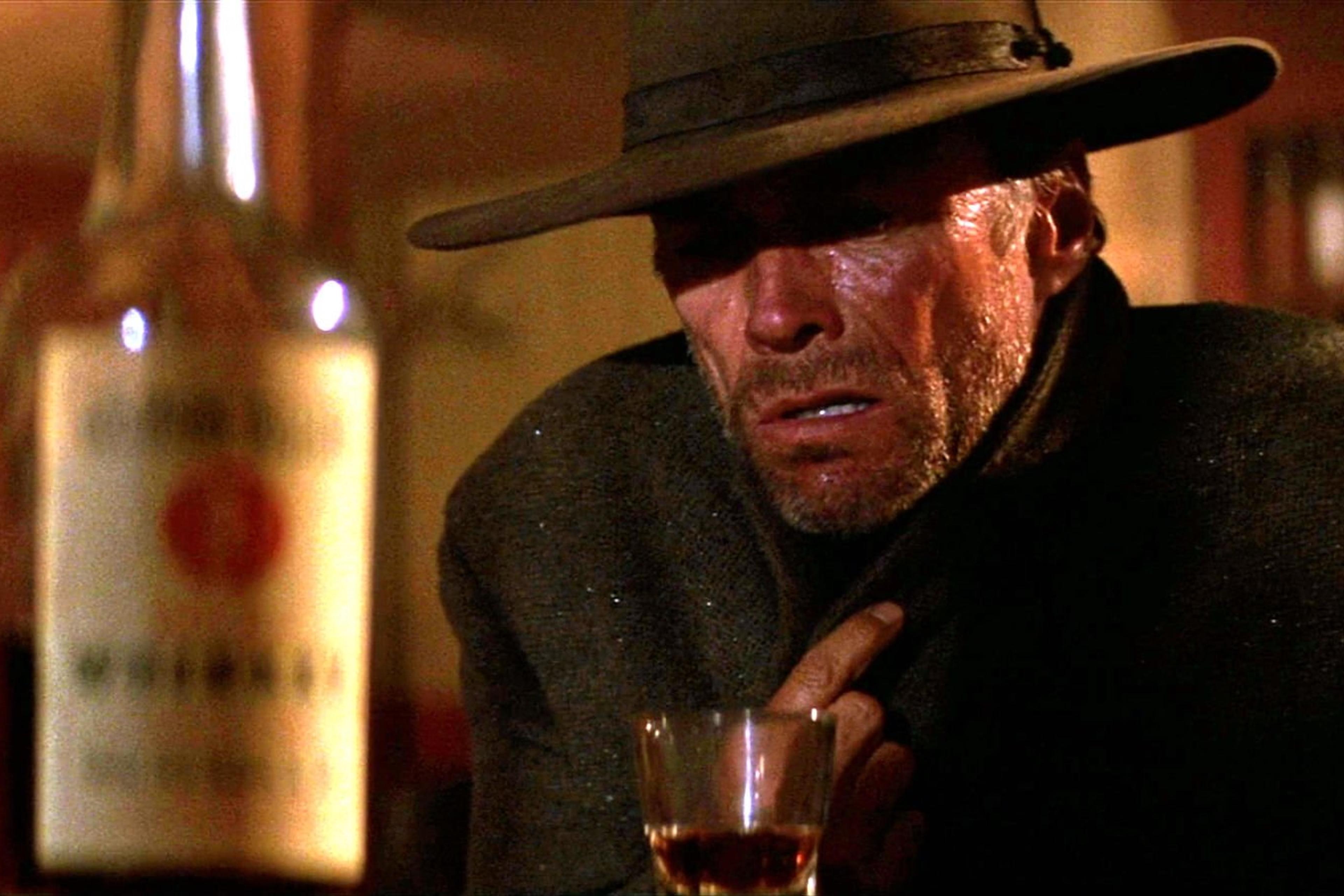 Scene from a movie with a rugged man in a hat holding his collar, a glass and bottle of whisky on the table.