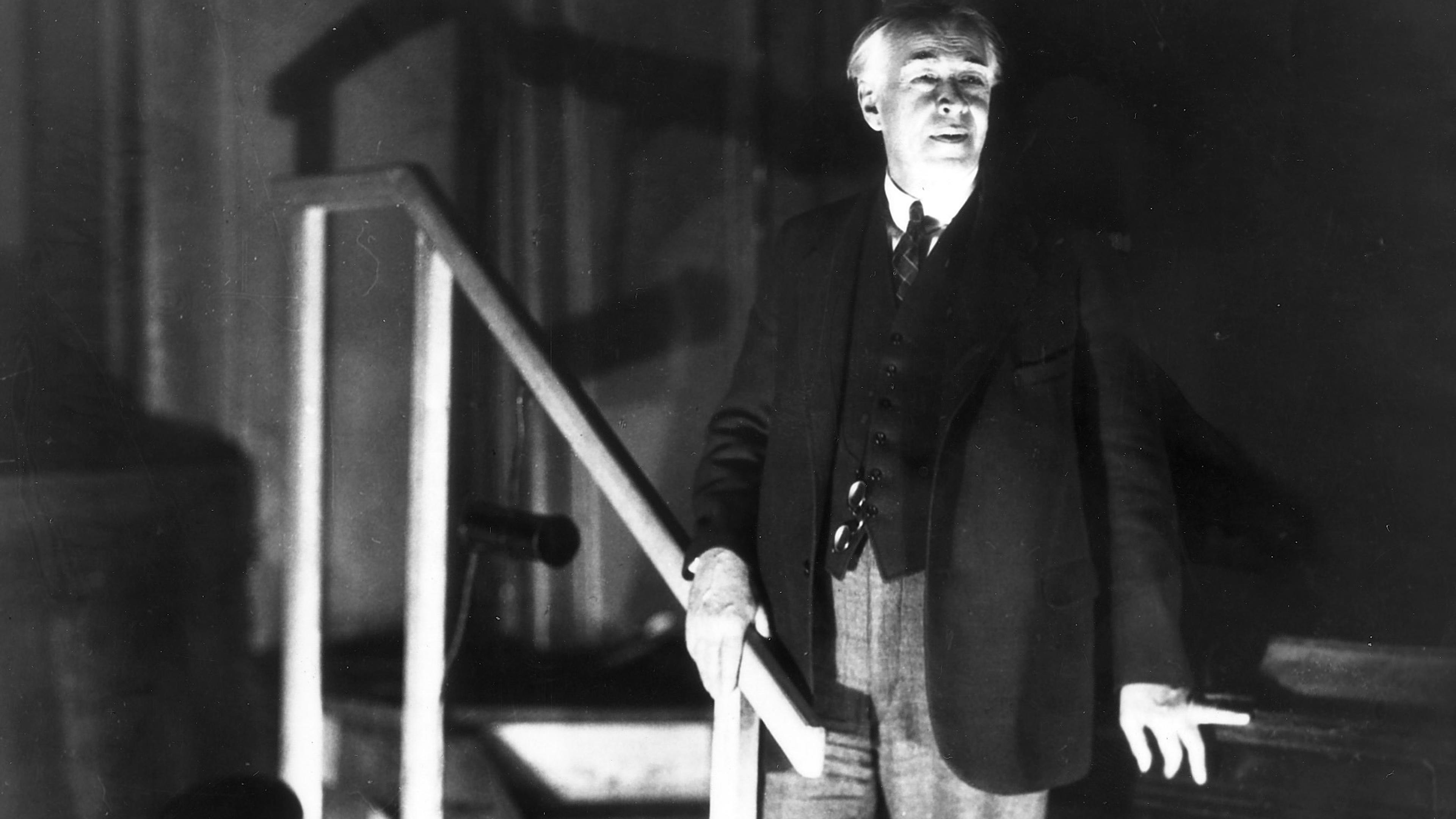 Black and white photo of a man in a suit standing by stairs in dramatic lighting.