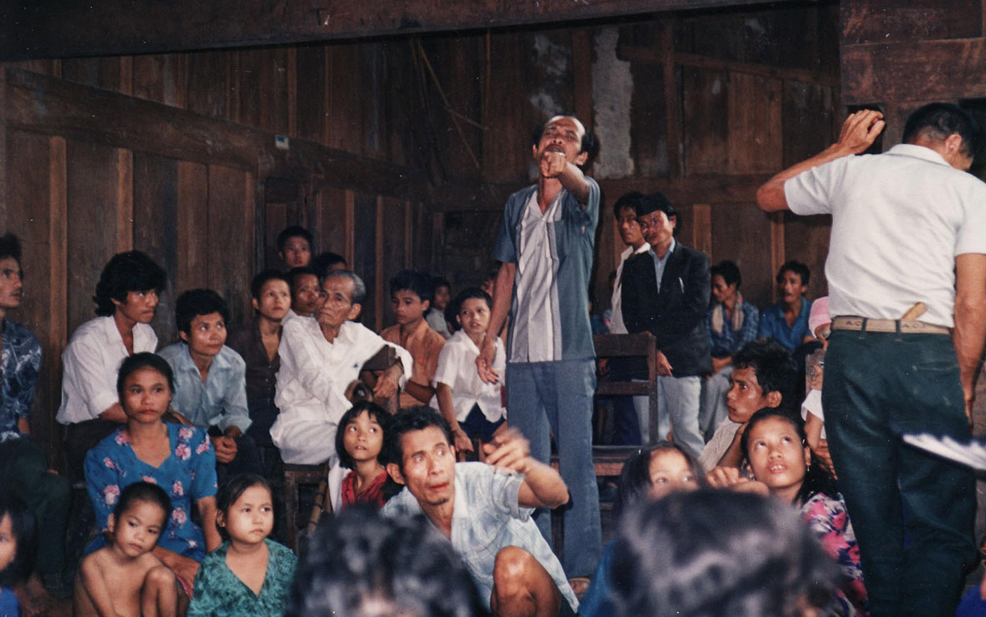 A group of Indonesian people sitting and standing in a wooden room. A man points while others, including children, look on with curiosity.