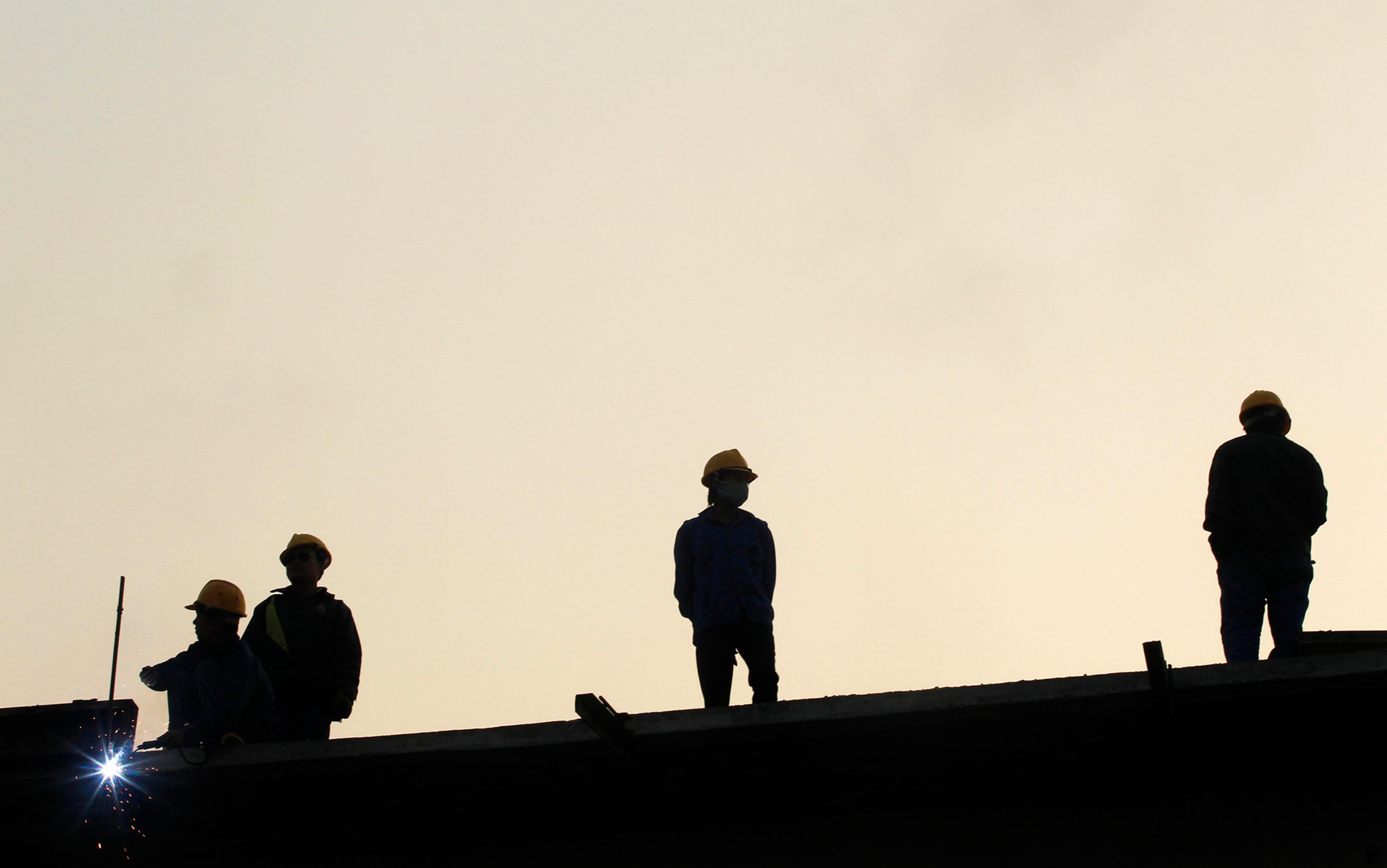 Silhouettes of four construction workers in hard hats standing on a beam against a sunset or sunrise sky. One worker is crouching and appearing to weld, emitting sparks.