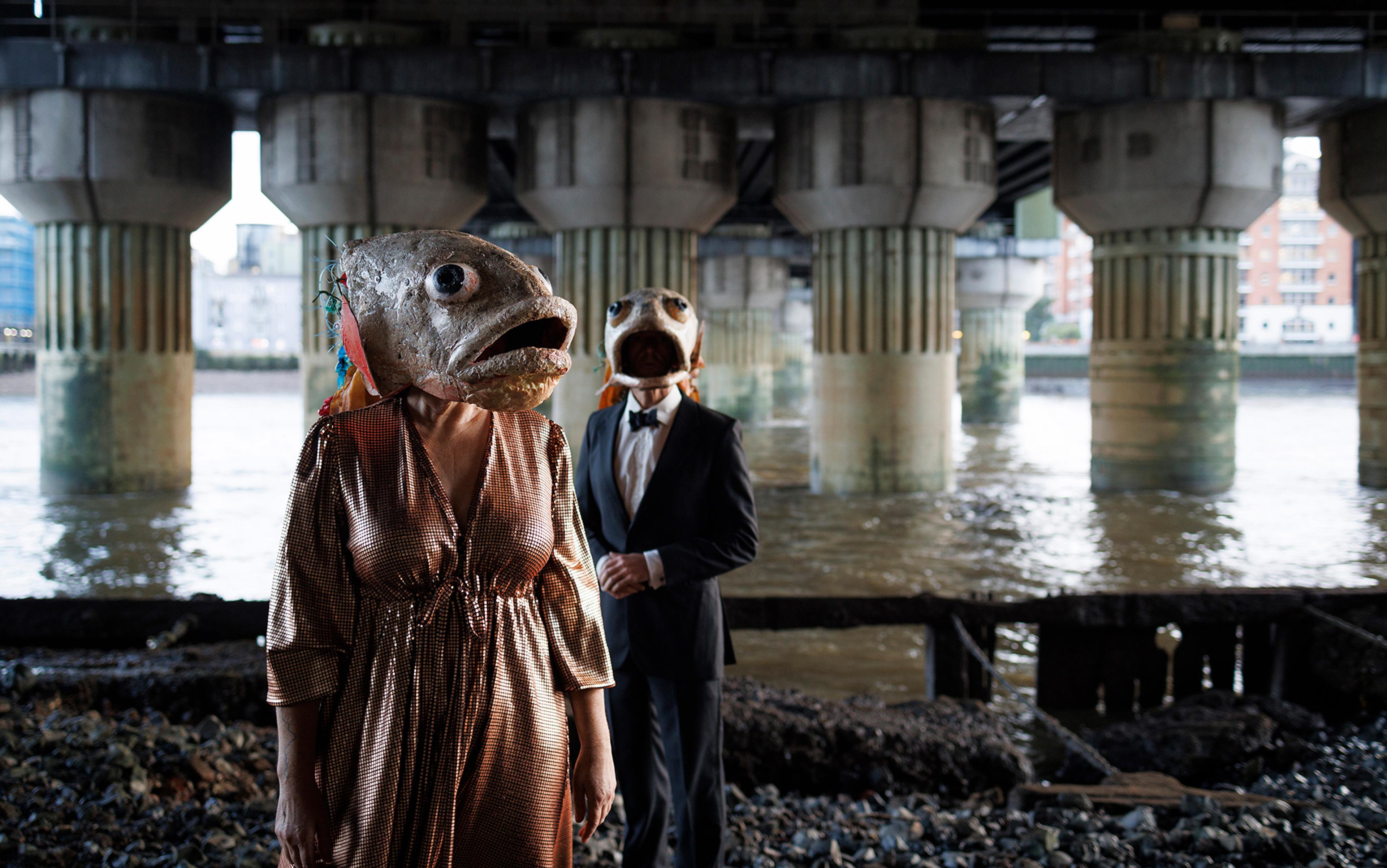 A man and a woman in formal evening dress but with giant fish heads covering their faces are pictured beneath a bridge on the foreshore of a river