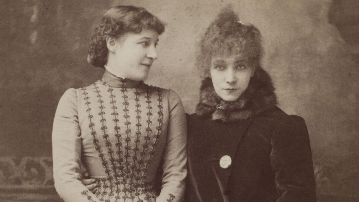 Vintage photo of two women in Victorian attire, one in a patterned dress looks at the other who wears a fur-trimmed coat.
