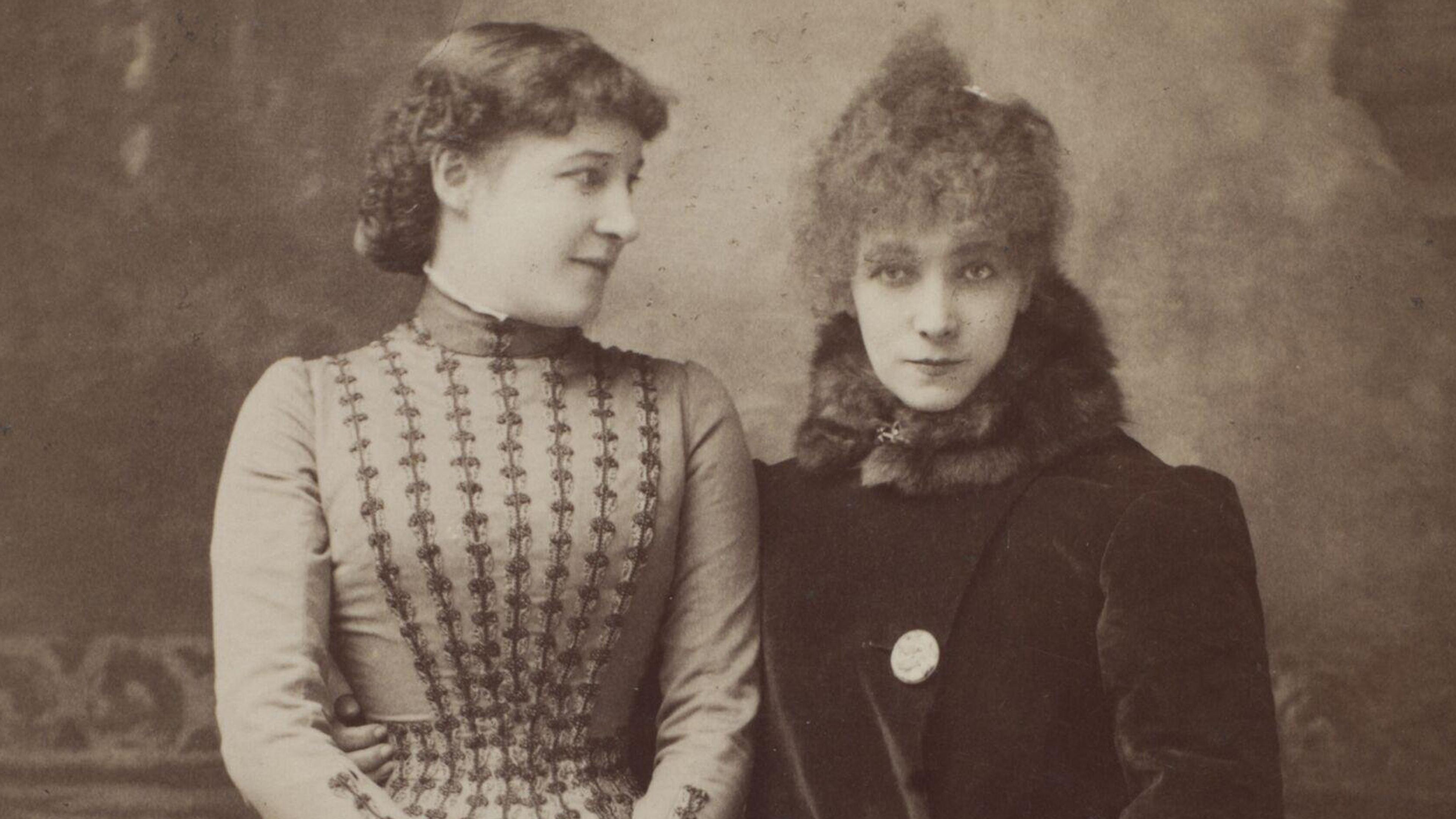 Vintage photo of two women in Victorian attire, one in a patterned dress looks at the other who wears a fur-trimmed coat.