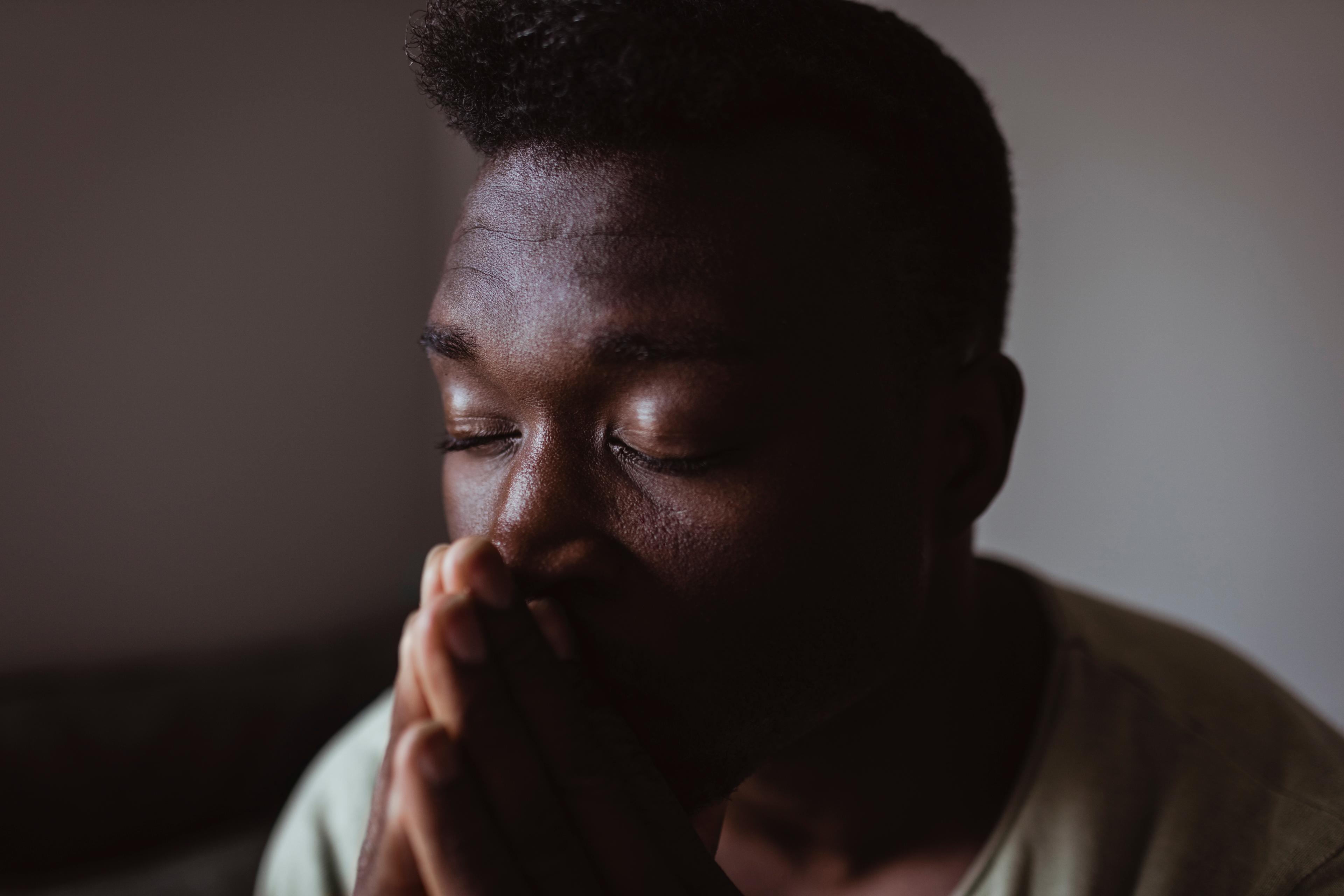Photo of a person with closed eyes and hands clasped, appearing thoughtful or in prayer, set against a dim background.