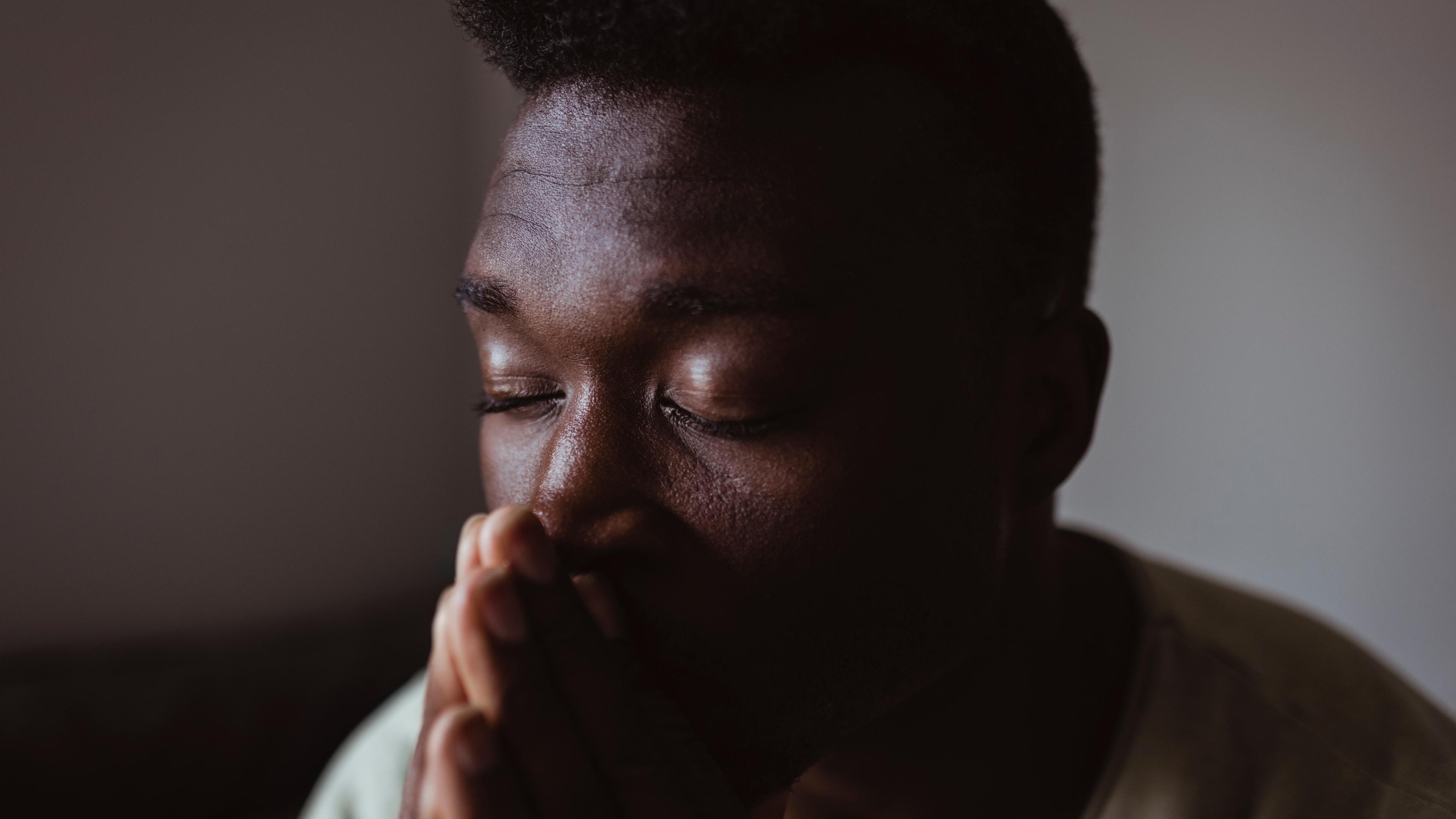 Photo of a person with closed eyes and hands clasped, appearing thoughtful or in prayer, set against a dim background.