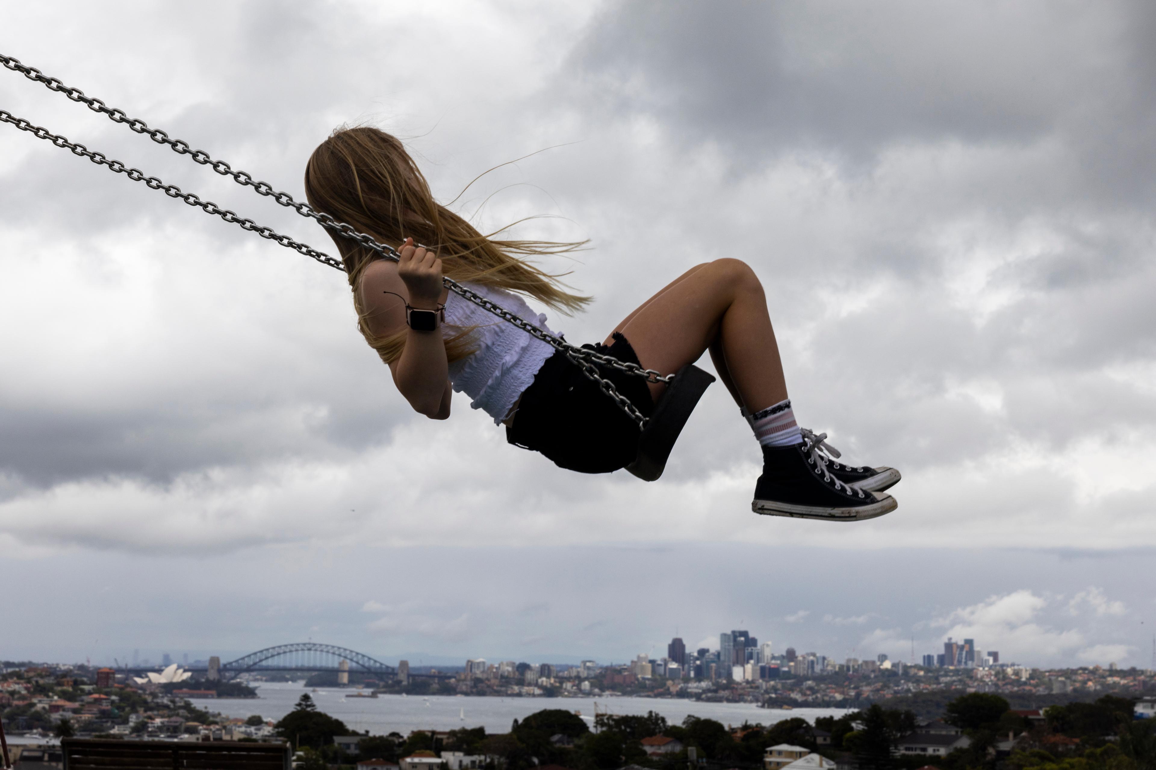 Photo of a girl swinging high above a cityscape with a cloudy sky in the background.