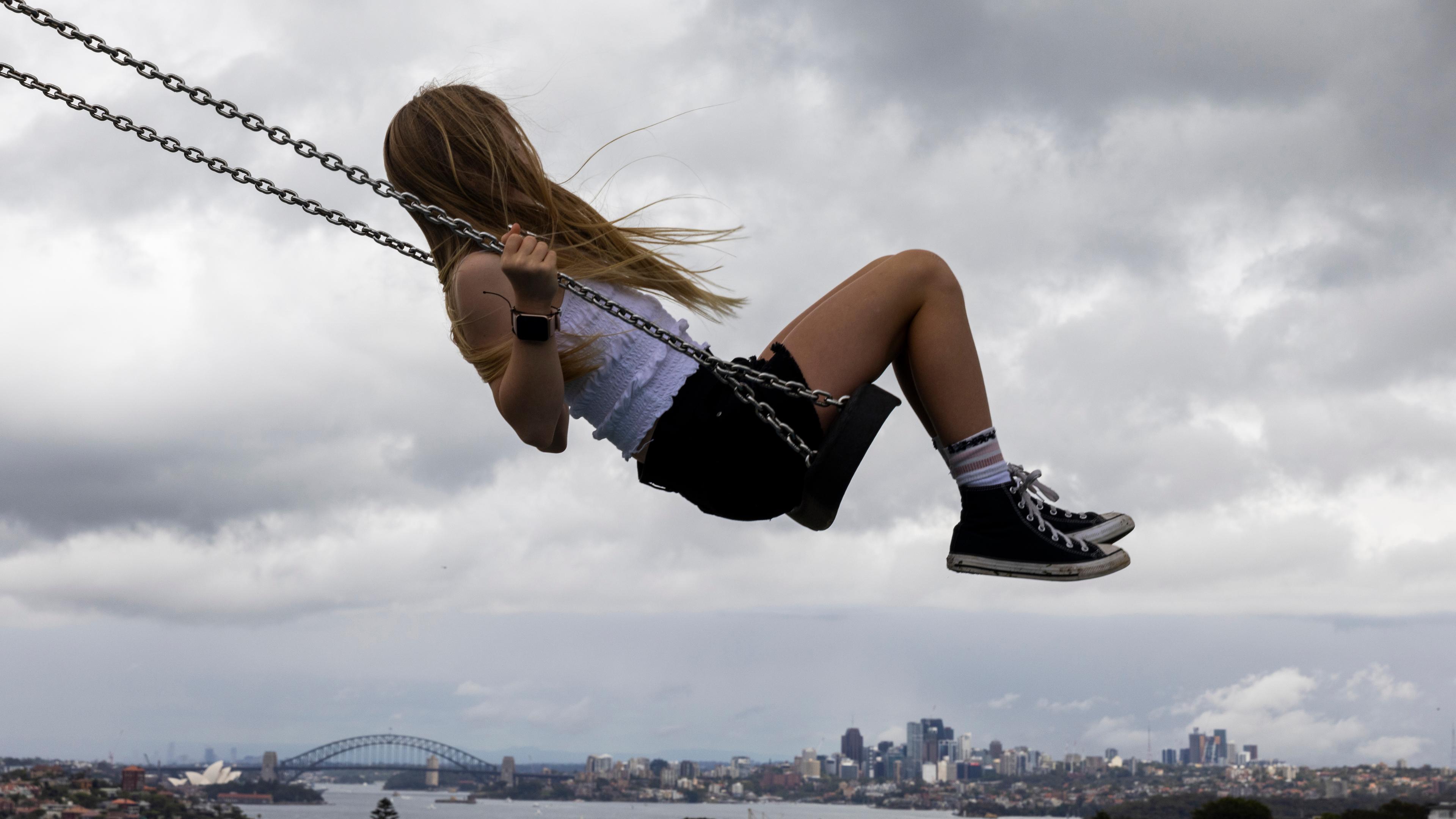 Photo of a girl swinging high above a cityscape with a cloudy sky in the background.