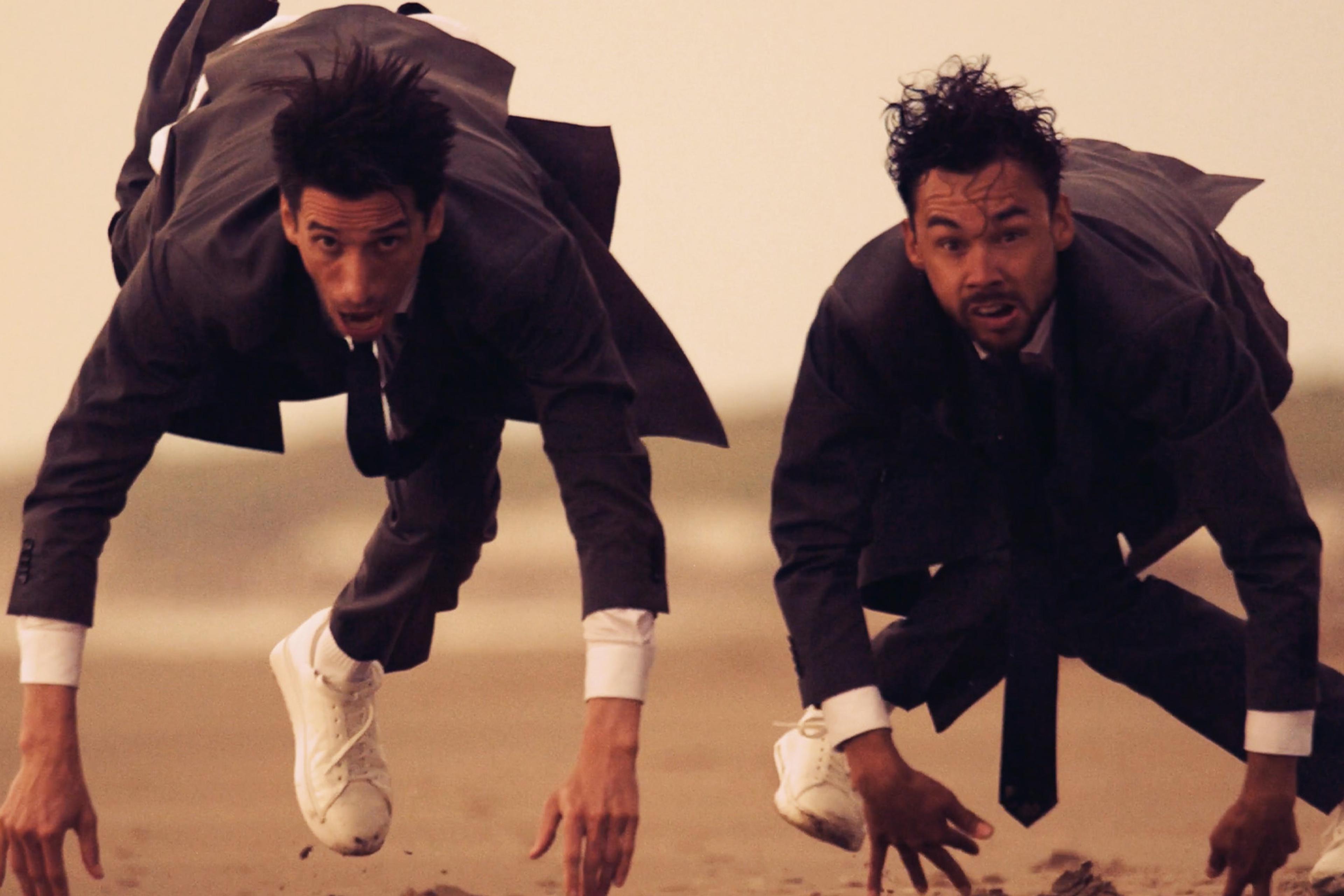 Two men in suits and white trainers crouch on a sandy beach, appearing to sprint forward with intense expressions.