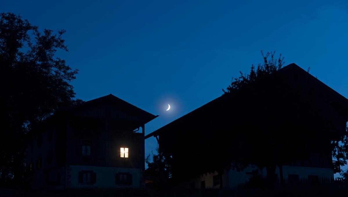 Houses at night with a crescent moon in a clear blue sky, one house has an illuminated window, tree silhouettes frame the scene.