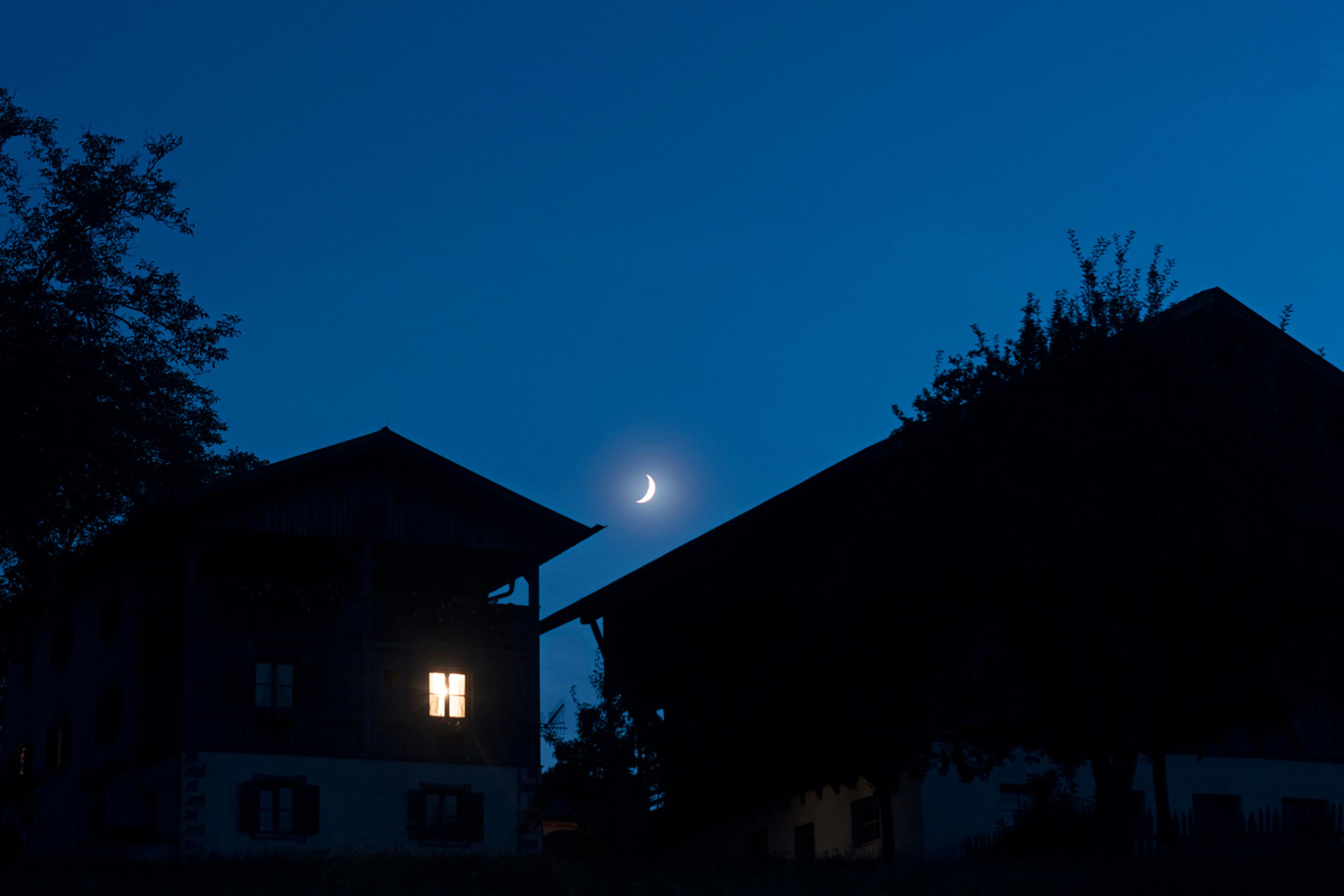 Houses at night with a crescent moon in a clear blue sky, one house has an illuminated window, tree silhouettes frame the scene.