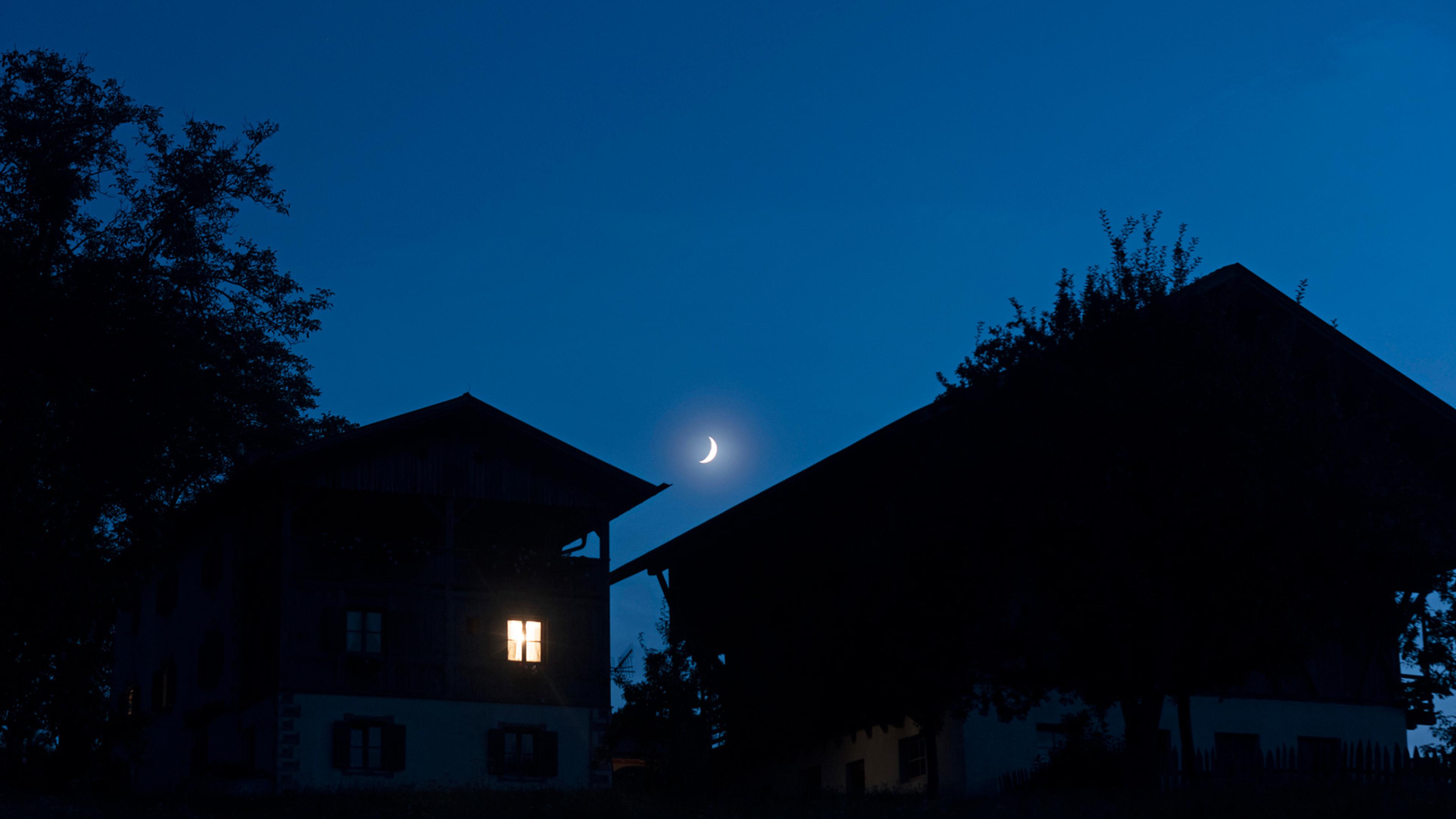 Houses at night with a crescent moon in a clear blue sky, one house has an illuminated window, tree silhouettes frame the scene.