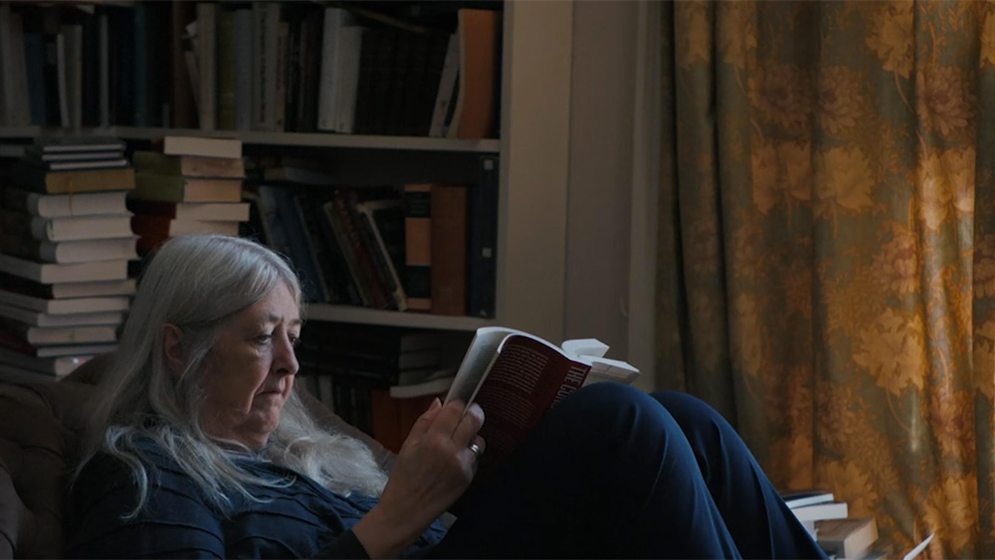 An older woman with grey hair sits with her knees pulled up and reads a book, seated by bookshelves, stacked books and a closed curtain.