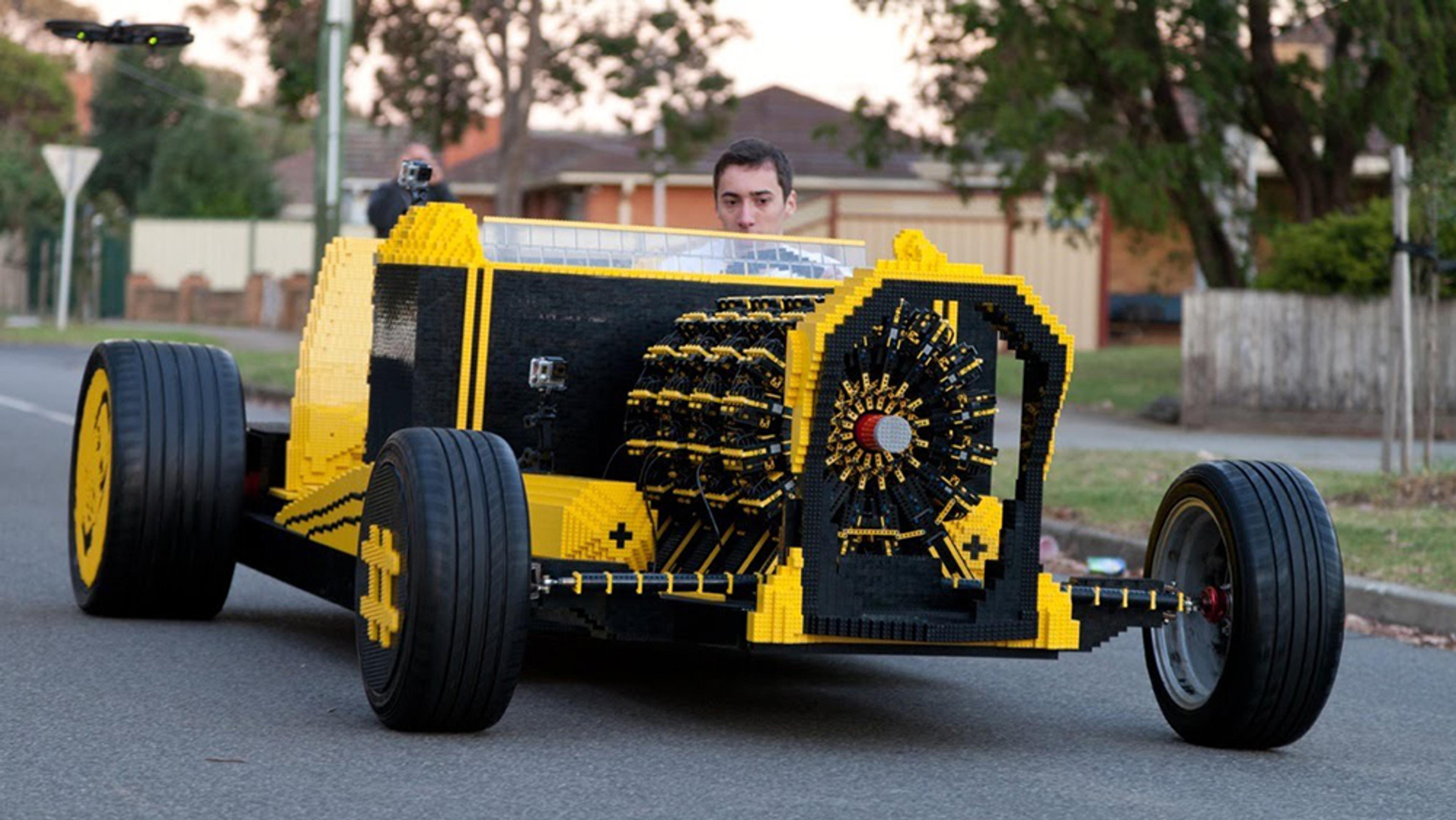 A person driving a life-sized car built from LEGO bricks on a suburban street with trees and houses in the background.