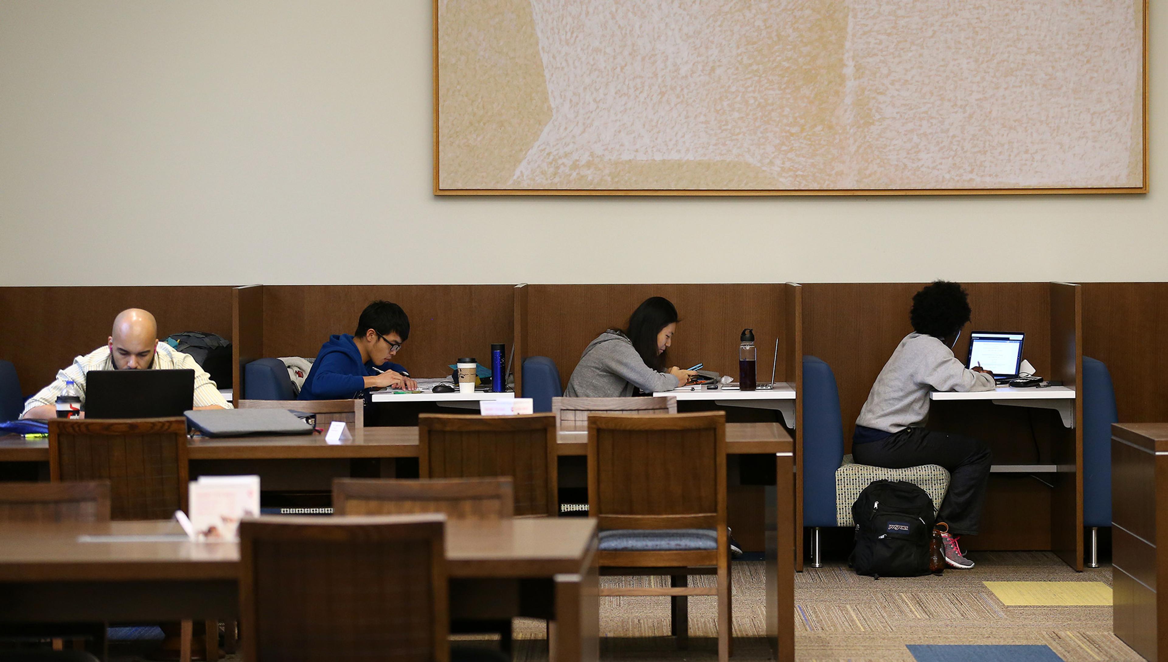 Students in a library, studying at their own private desk in cubicles or at a communal table