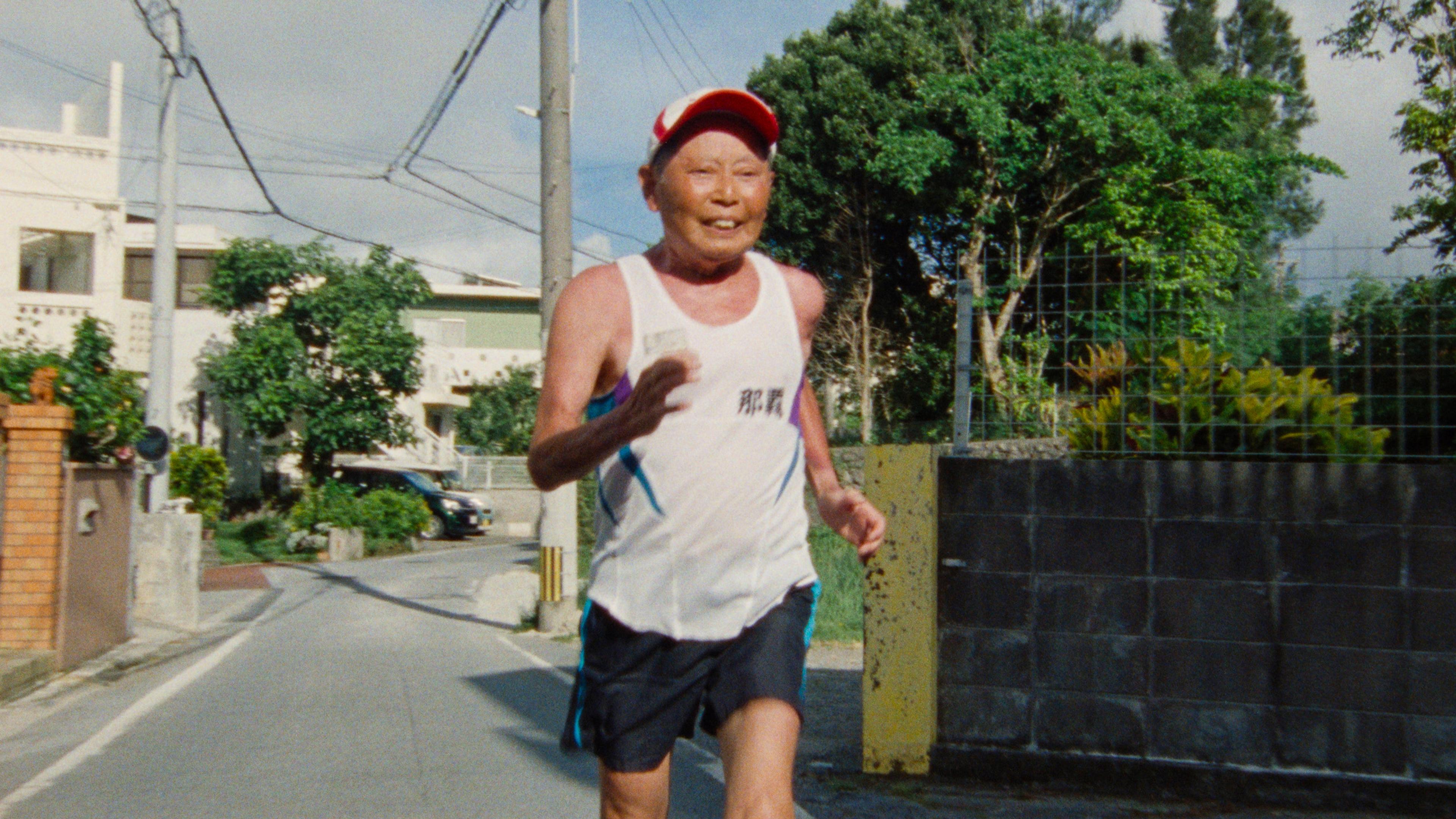 Photo of an elderly man jogging on a street lined with trees houses visible in the background on a clear day.