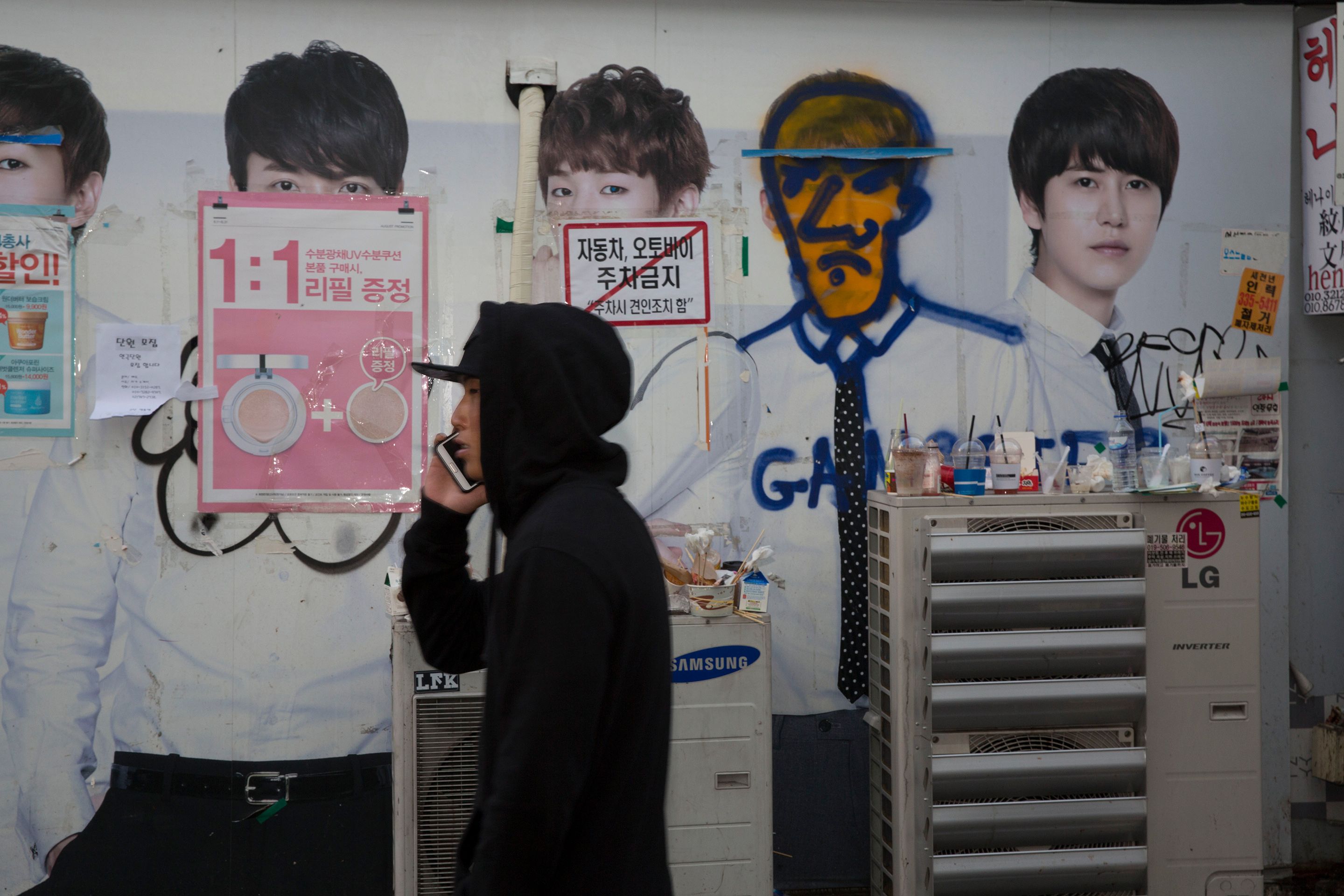 A man in a hoodie on a phone walking past a wall with posters and graffiti-covered advertisements.