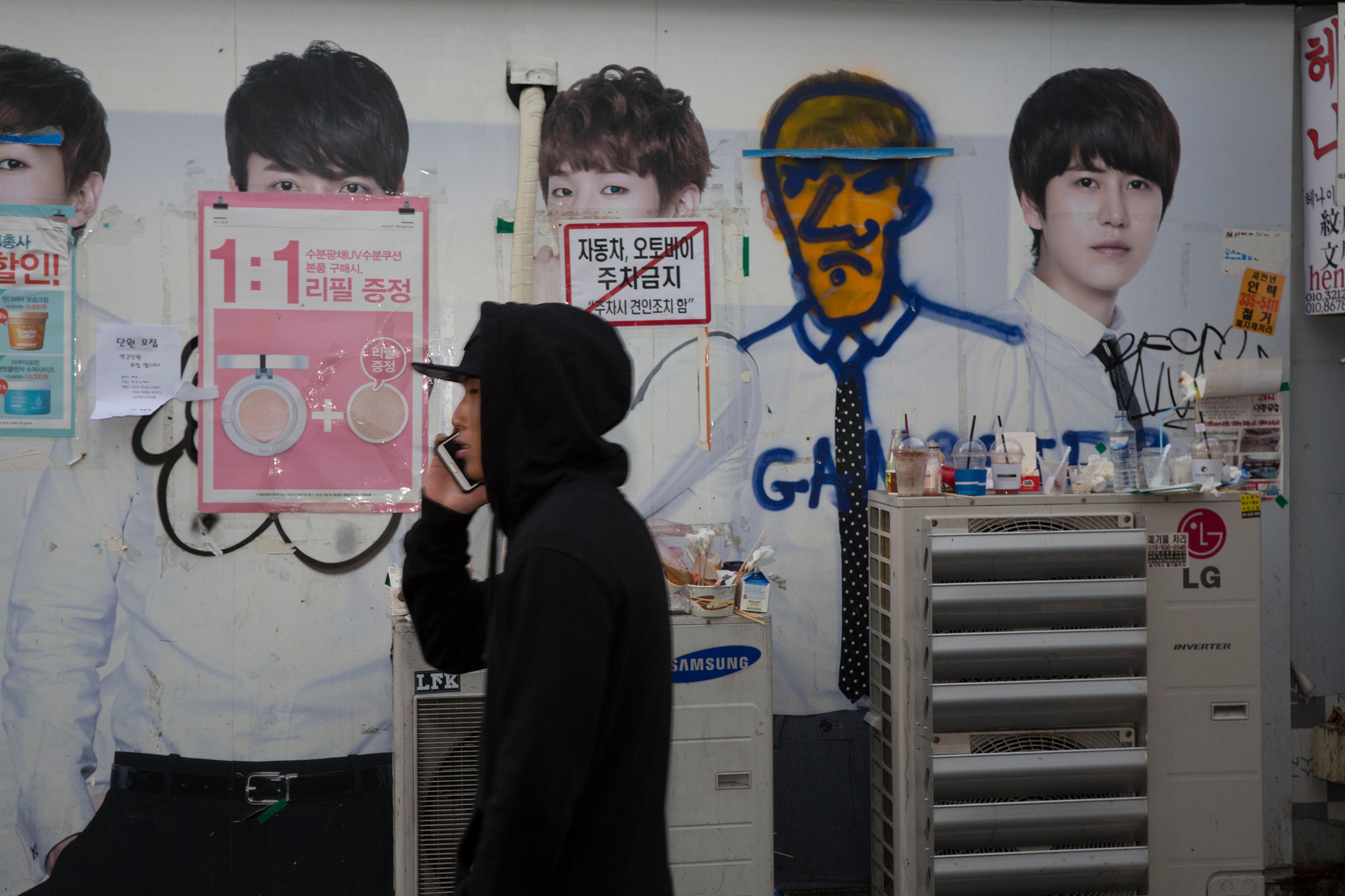 A man in a hoodie on a phone walking past a wall with posters and graffiti-covered advertisements.