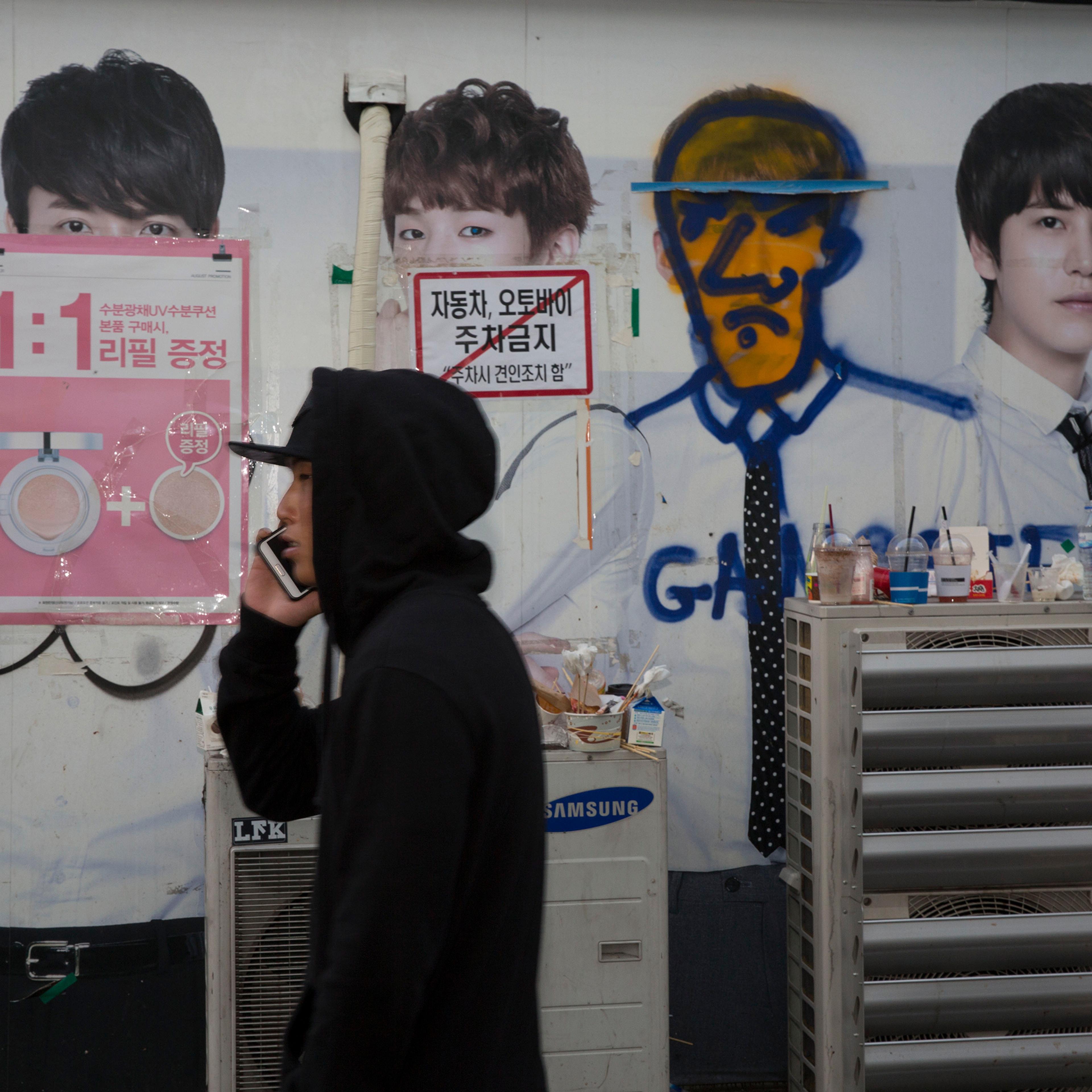 A man in a hoodie on a phone walking past a wall with posters and graffiti-covered advertisements.