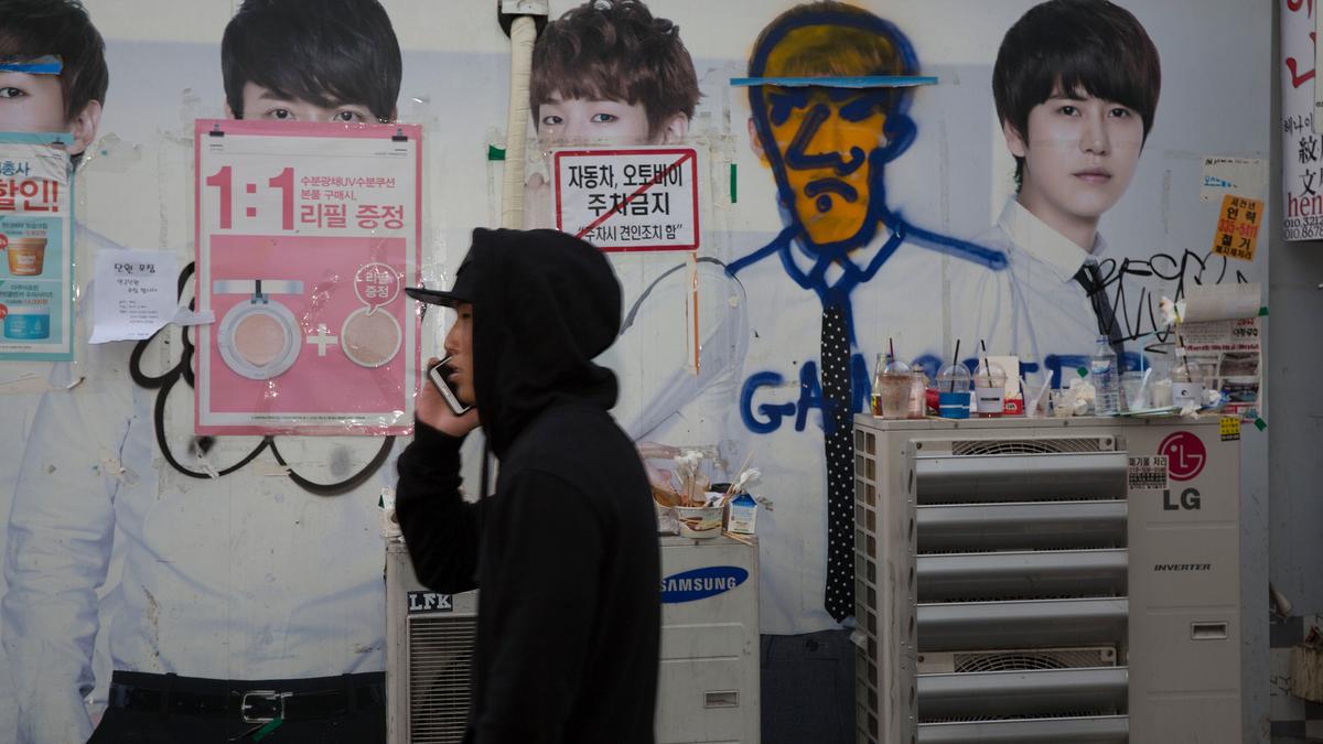 A man in a hoodie on a phone walking past a wall with posters and graffiti-covered advertisements.