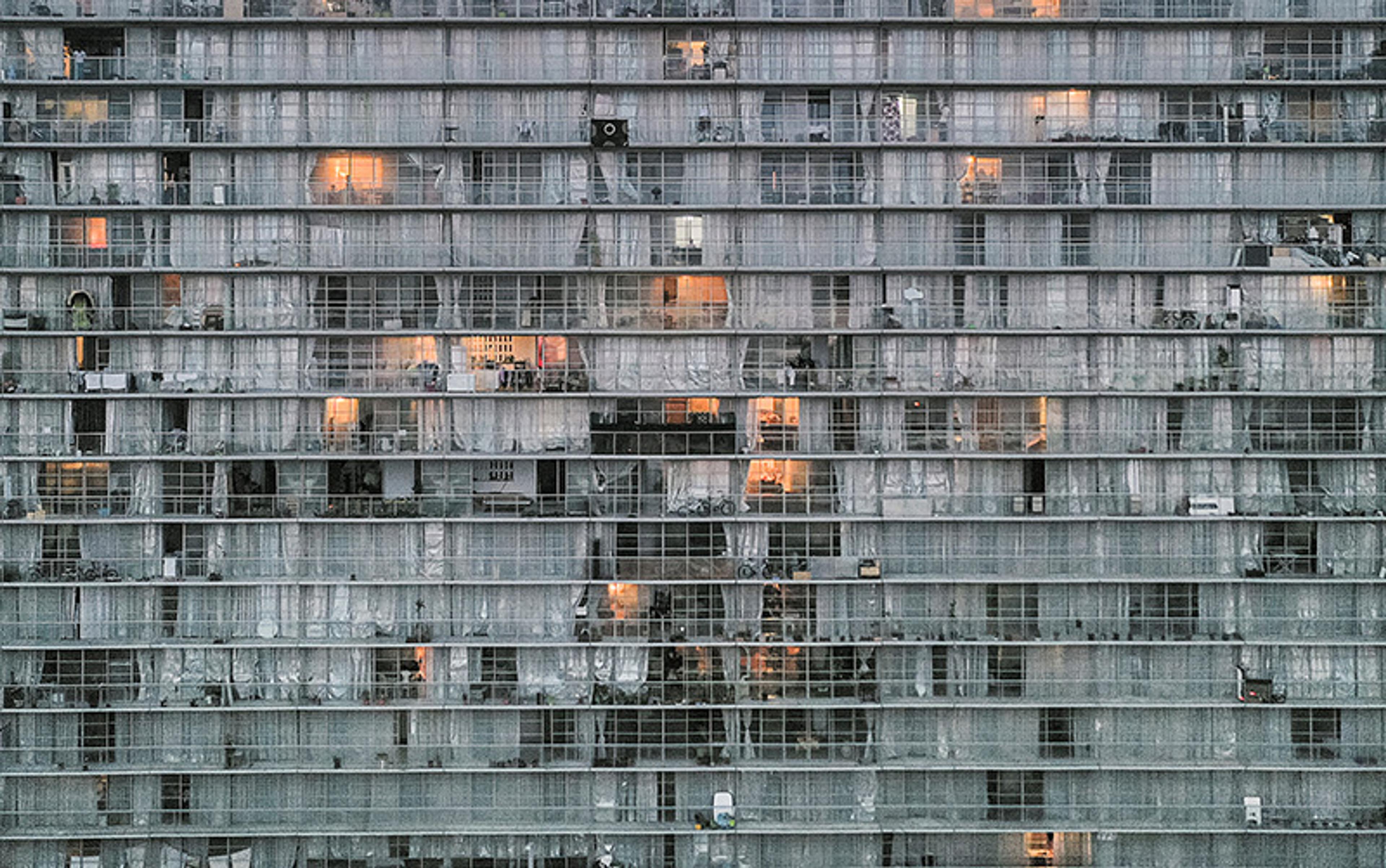 A high-rise building facade with rows of windows and balconies, some illuminated from within, creating a grid-like pattern.