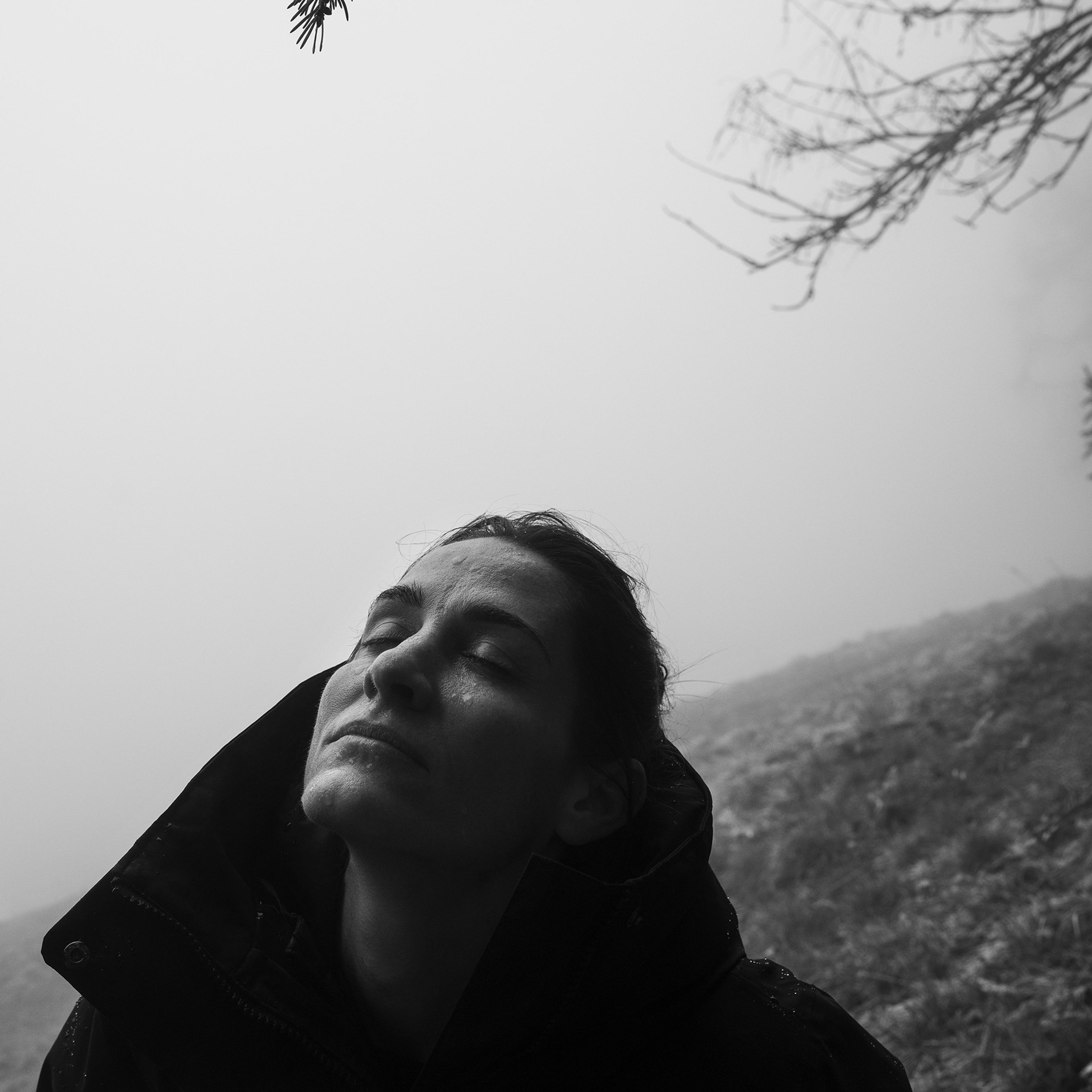 Black-and-white photograph of a person in a jacket standing outdoors in a foggy environment, looking up with eyes closed.