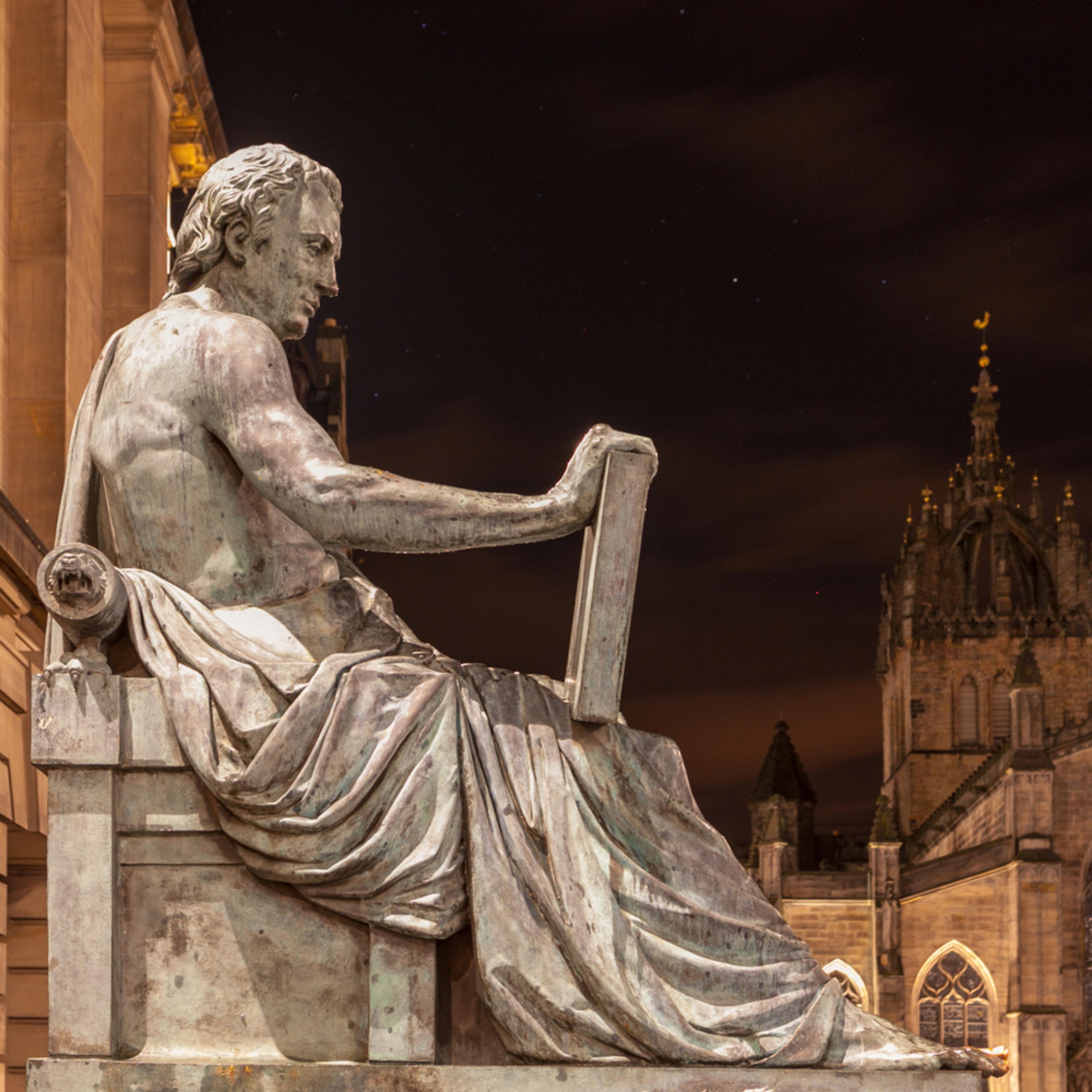 A statue of a seated man with a tablet at night with buildings in the background, featuring detailed drapery and classical design.