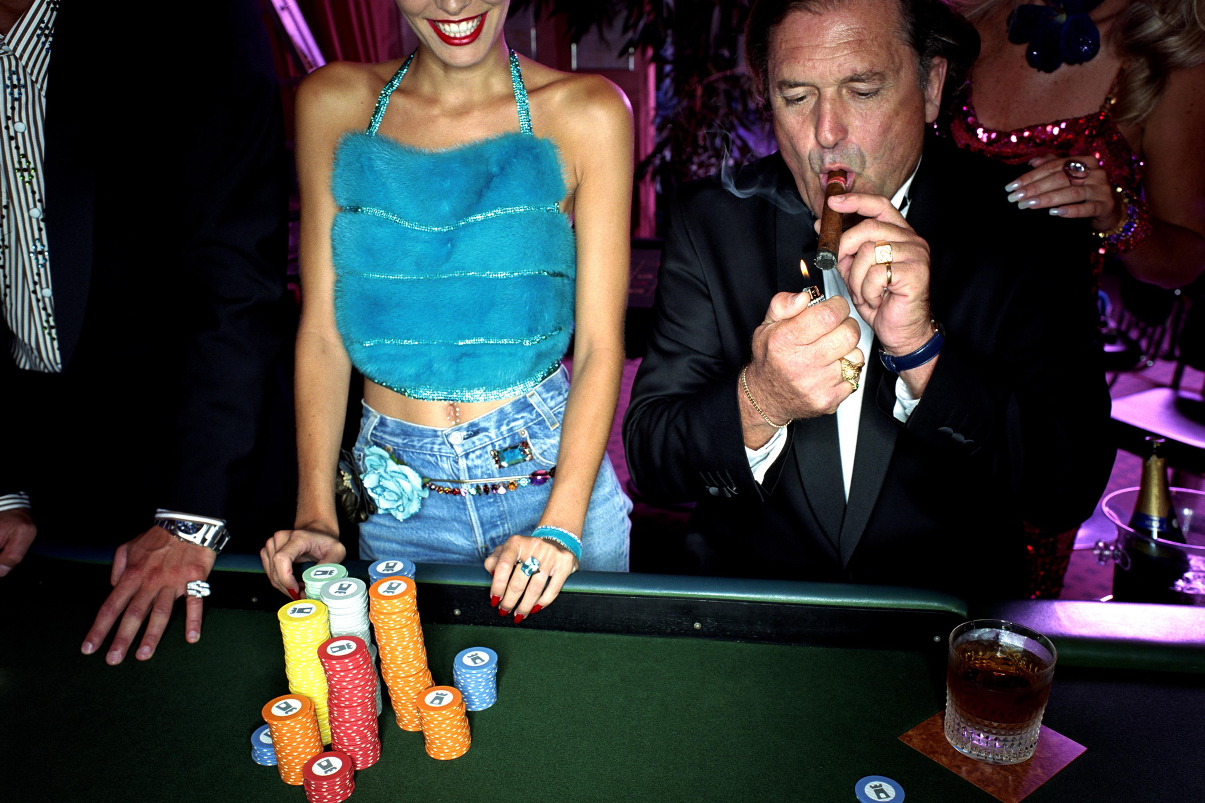 Photo of a casino scene with people, poker chips on a table, and a man lighting a cigar. A woman in a blue top smiles.