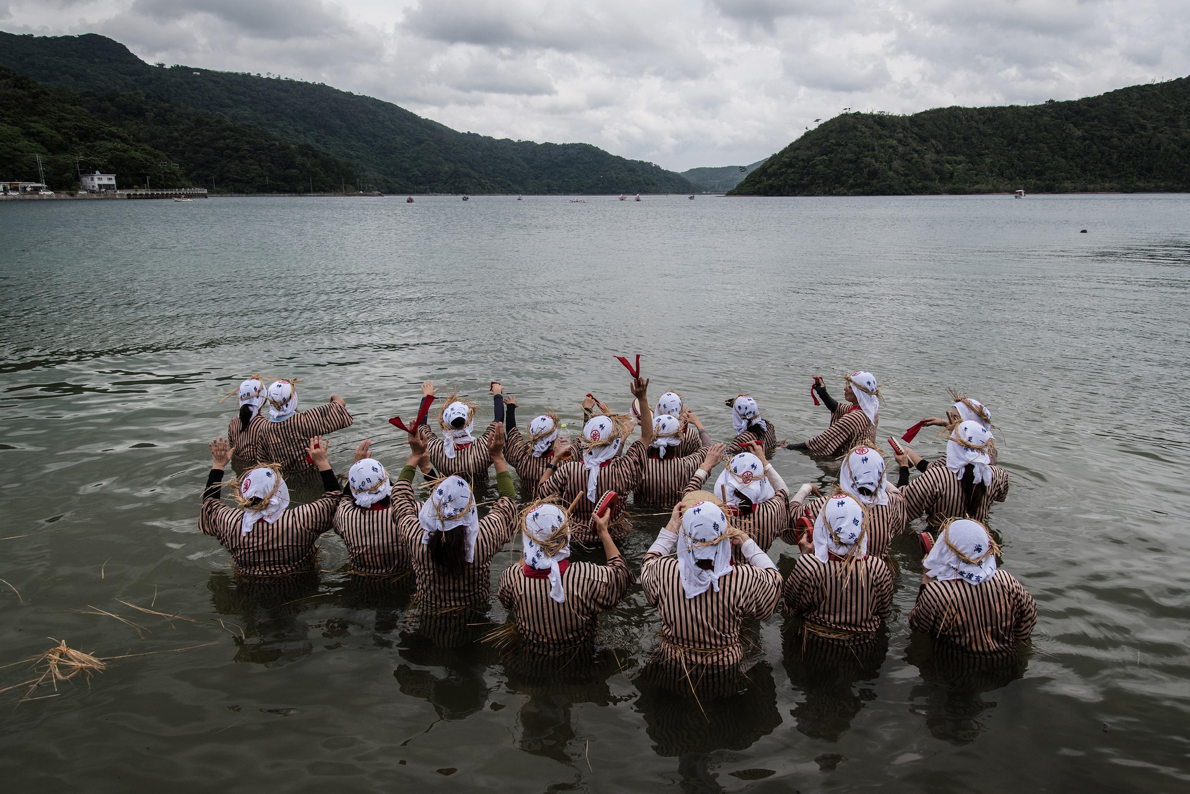 A group of people in striped clothing and headscarves standing in shallow water, raising hands towards a cloudy sky.
