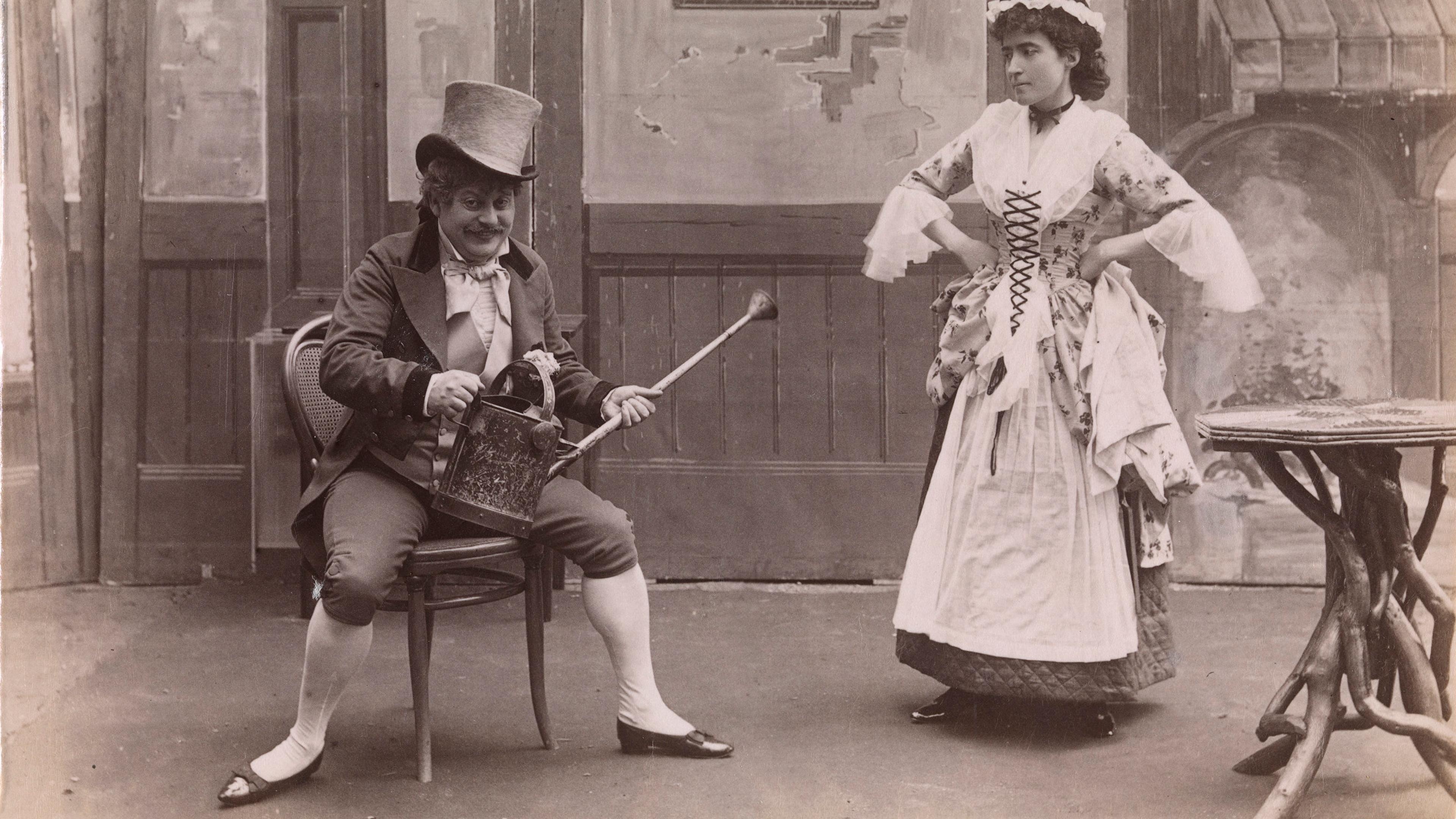 Black and white photo of a man in a top hat with a watering can seated, woman standing in period attire, vintage interior.
