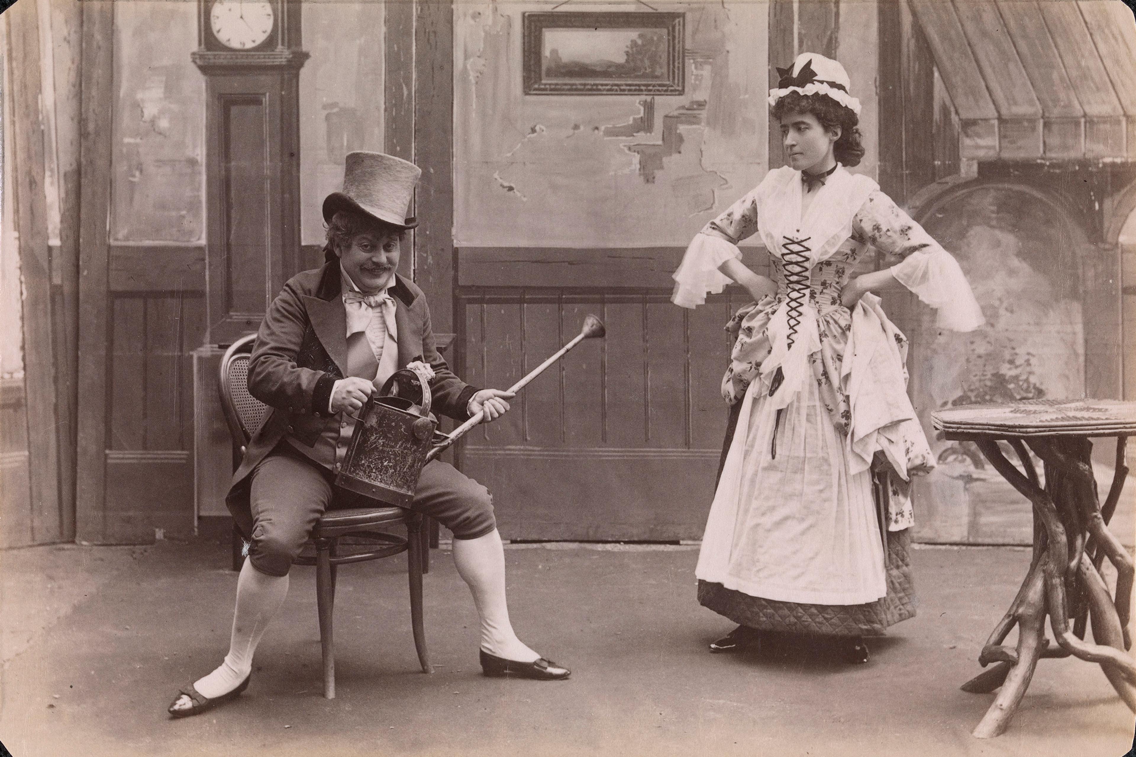 Black and white photo of a man in a top hat with a watering can seated, woman standing in period attire, vintage interior.
