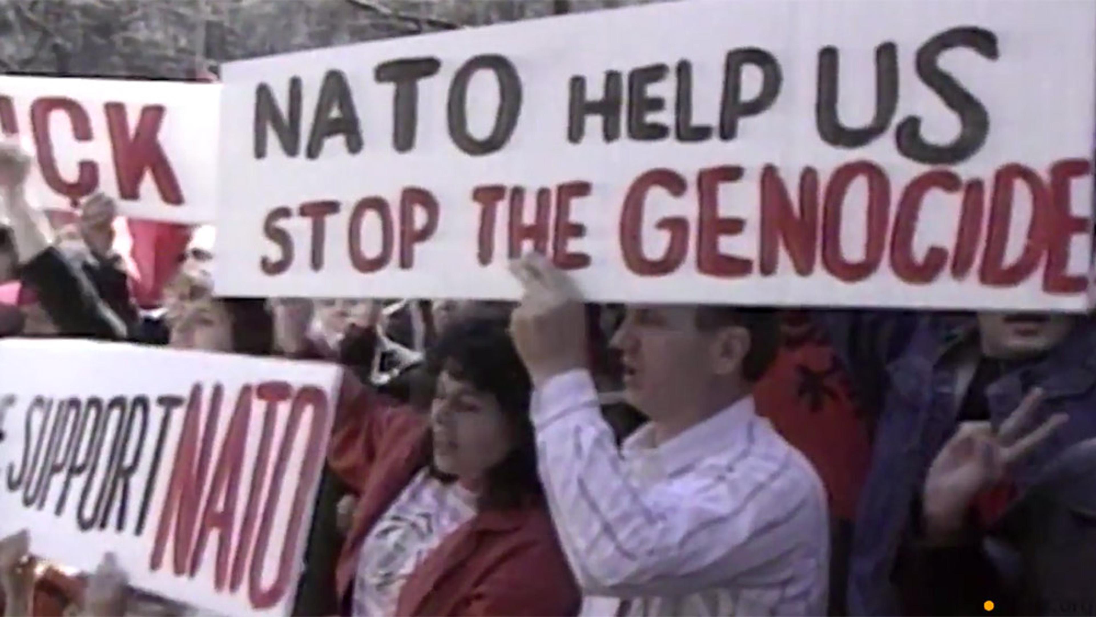 Photo of a group of people holding signs that read “NATO help us stop the genocide” and “We support NATO” at a demonstration.