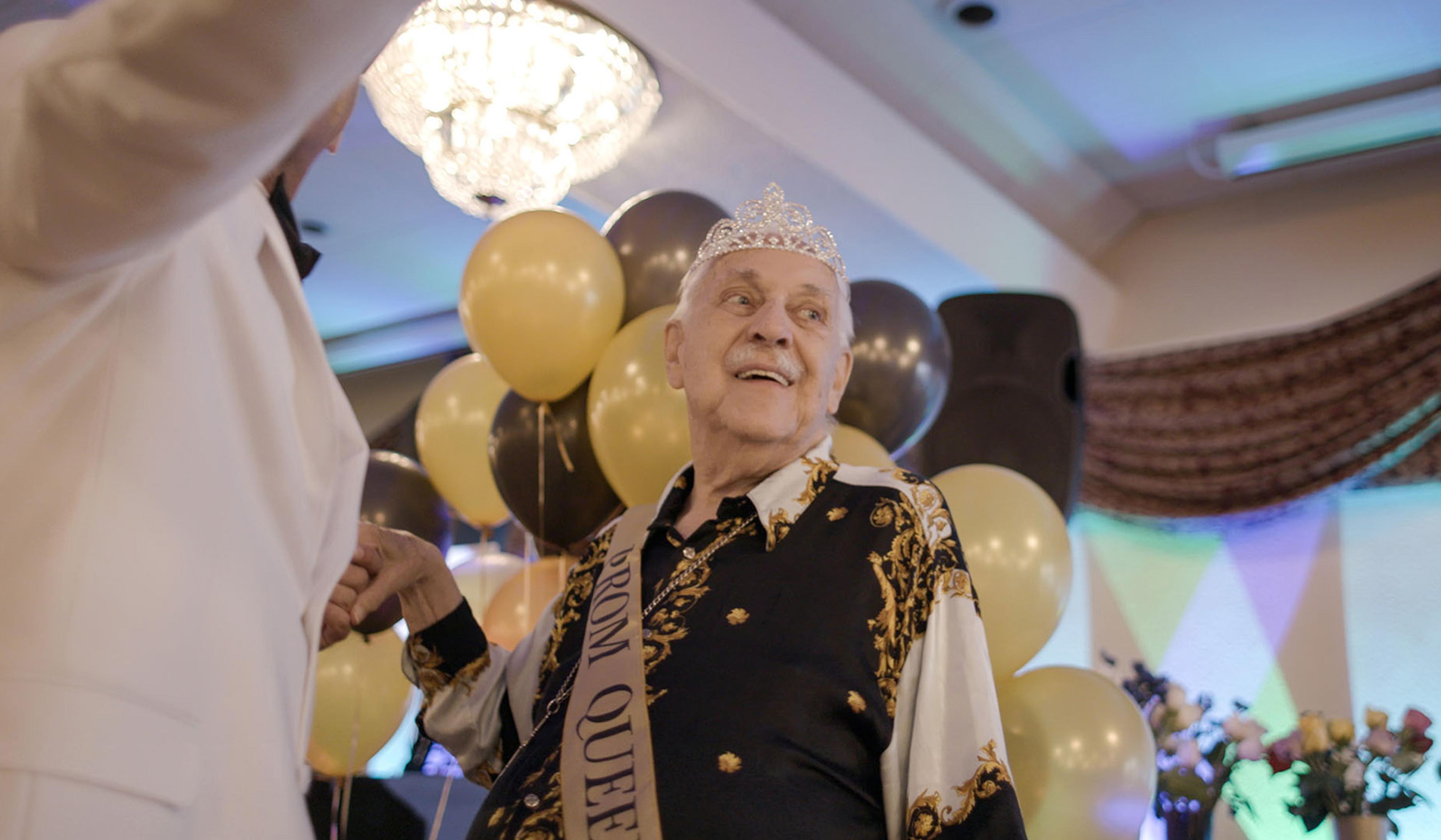 Elderly person wearing a tiara and a “Prom Queen” sash, smiling at a dance, with balloons and a chandelier in the background.