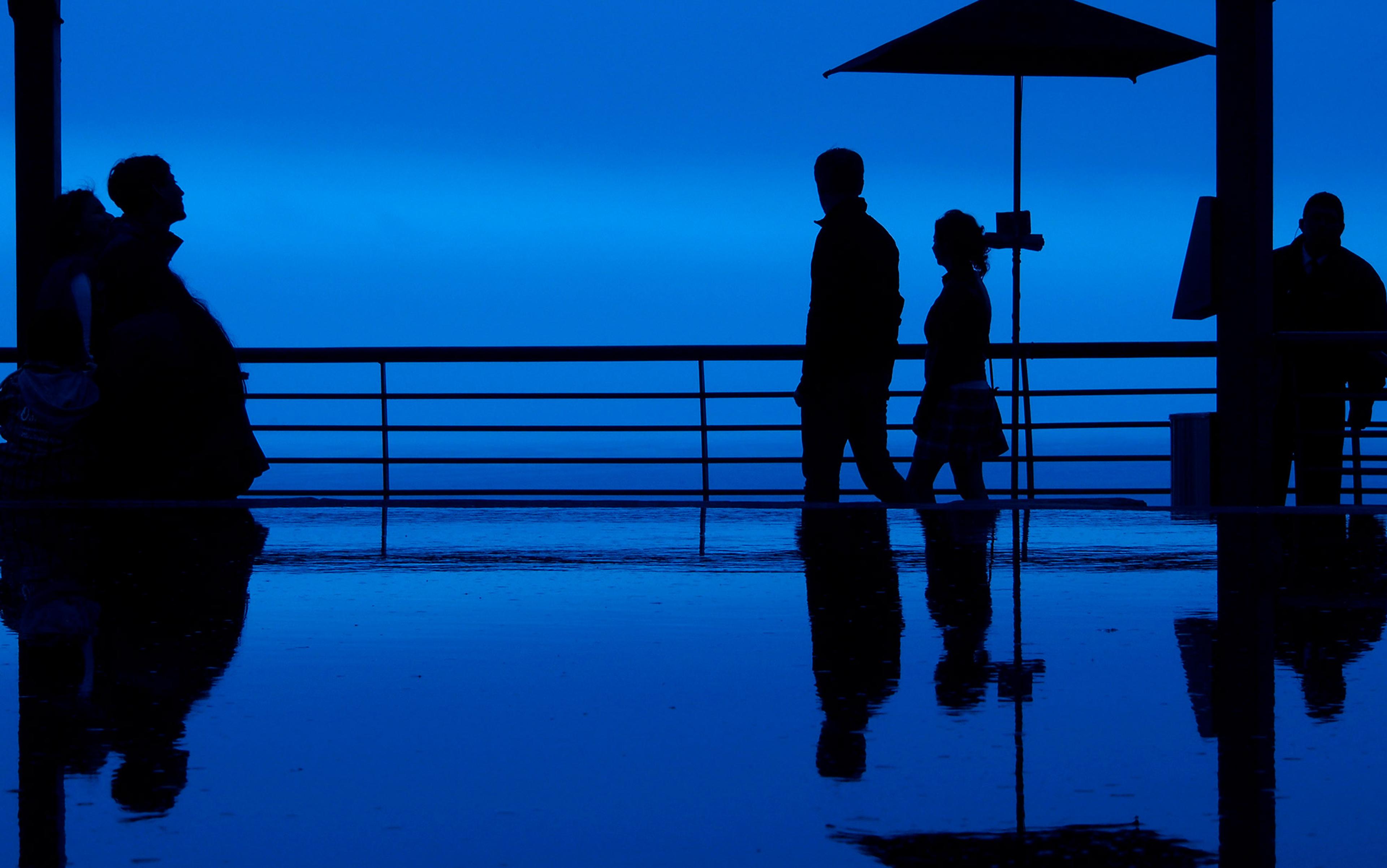 Silhouettes of people walking and standing by a railing against a blue evening sky, reflected in a wet surface below.