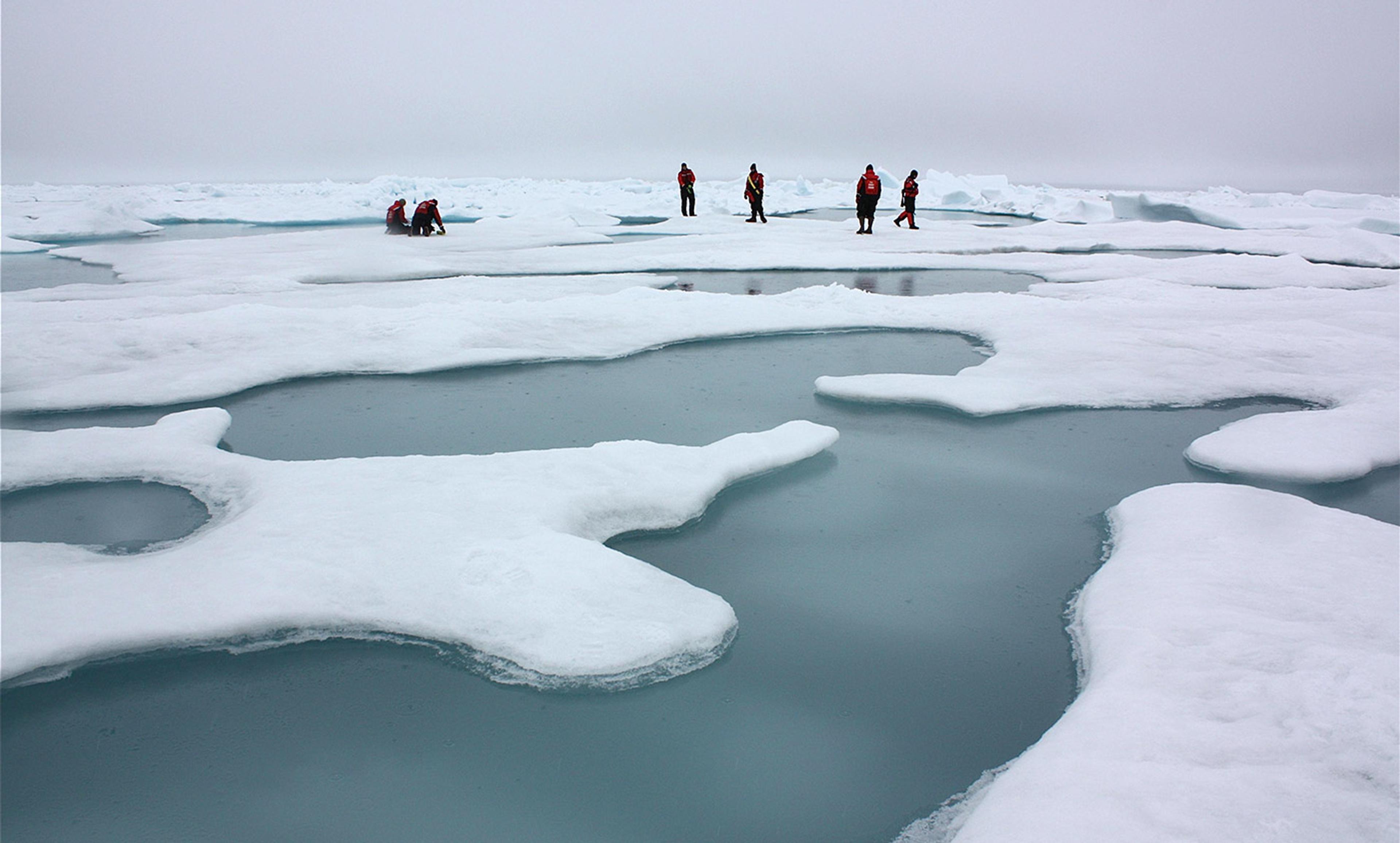 Several people in red jackets walking on an icy landscape with patches of open water under a grey sky.