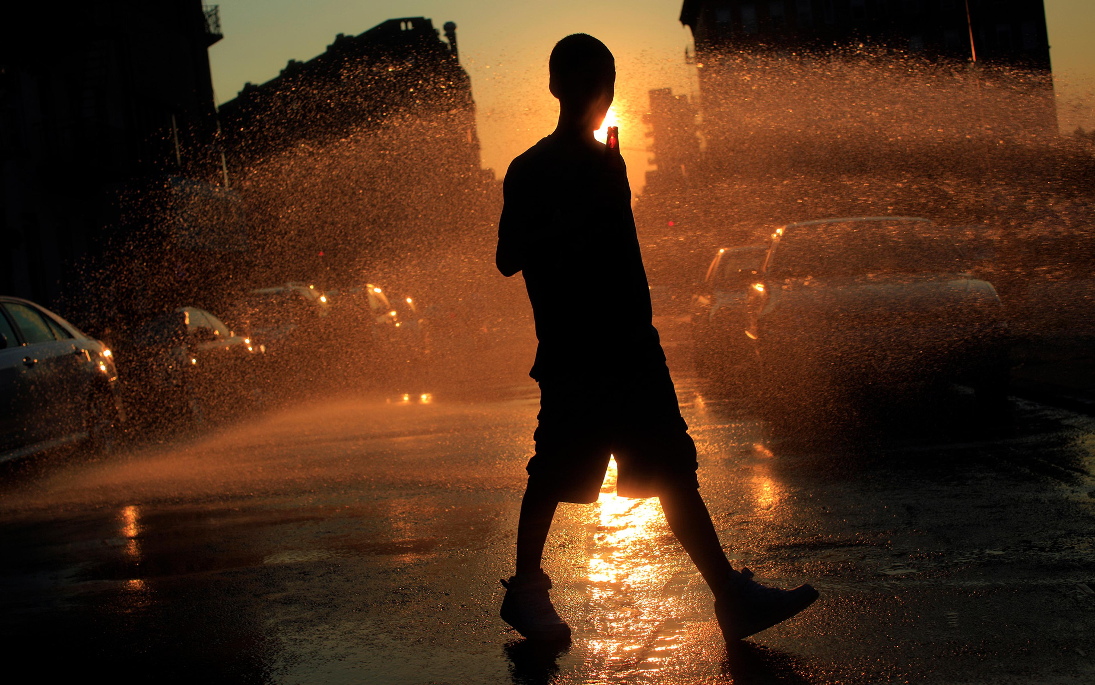 Silhouette of a person walking through a spray of water at sunset with cars and buildings in the background.