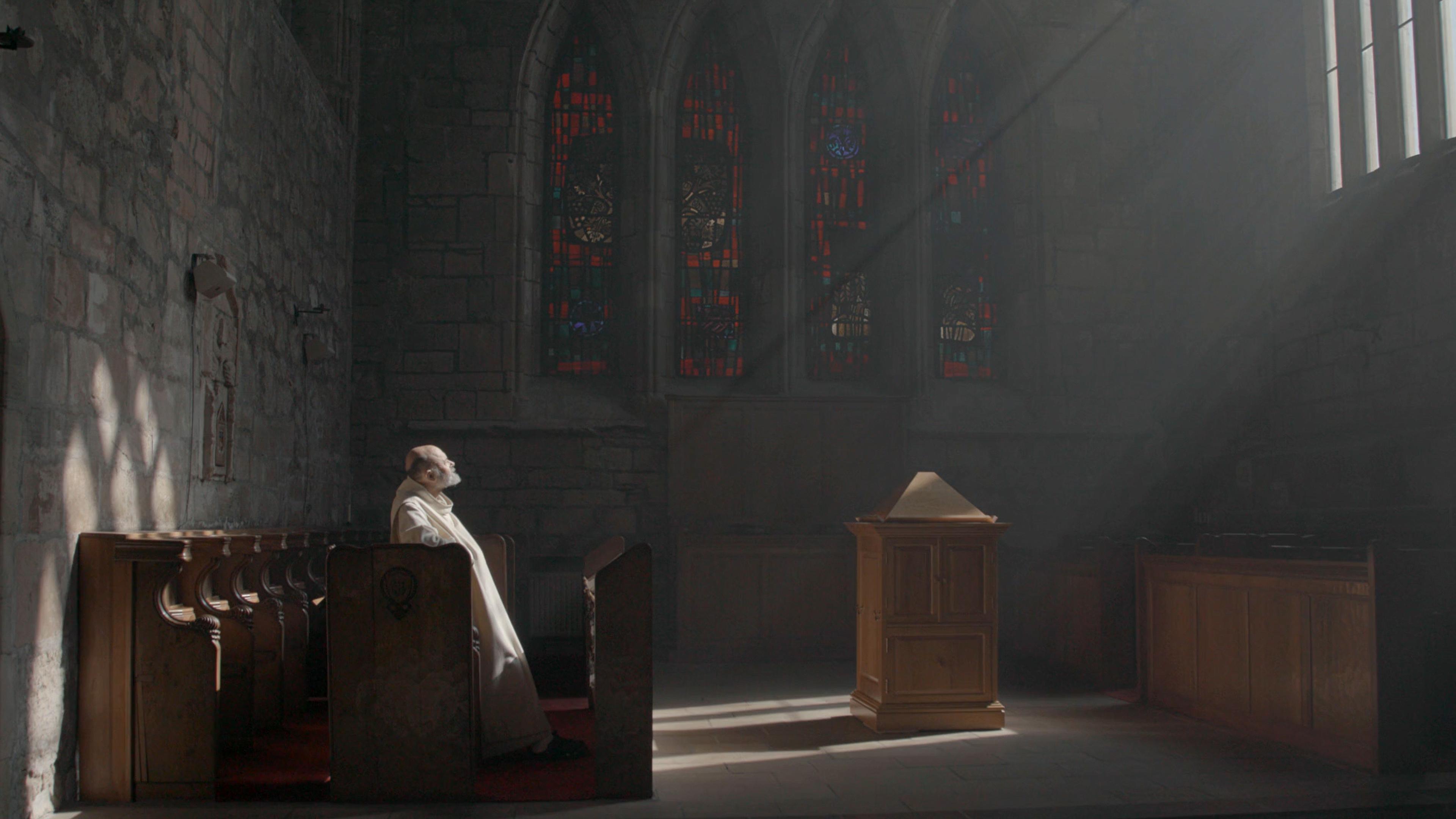 Photo of a person sitting in a dimly lit church with stained glass windows and sun rays filtering through.