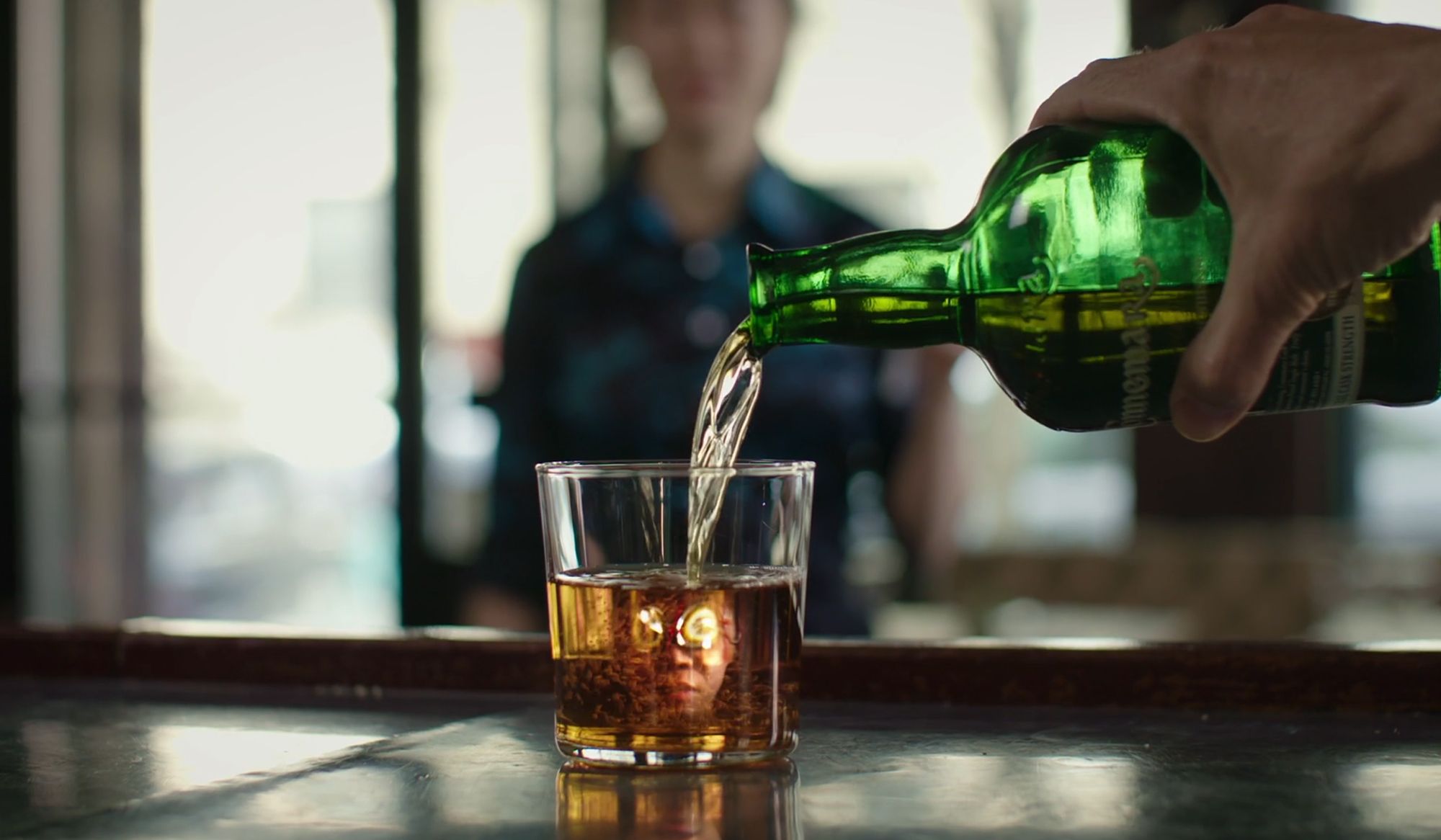 A hand pouring alcohol from a green bottle into a glass with a reflection of a face. Blurry background with a person standing.