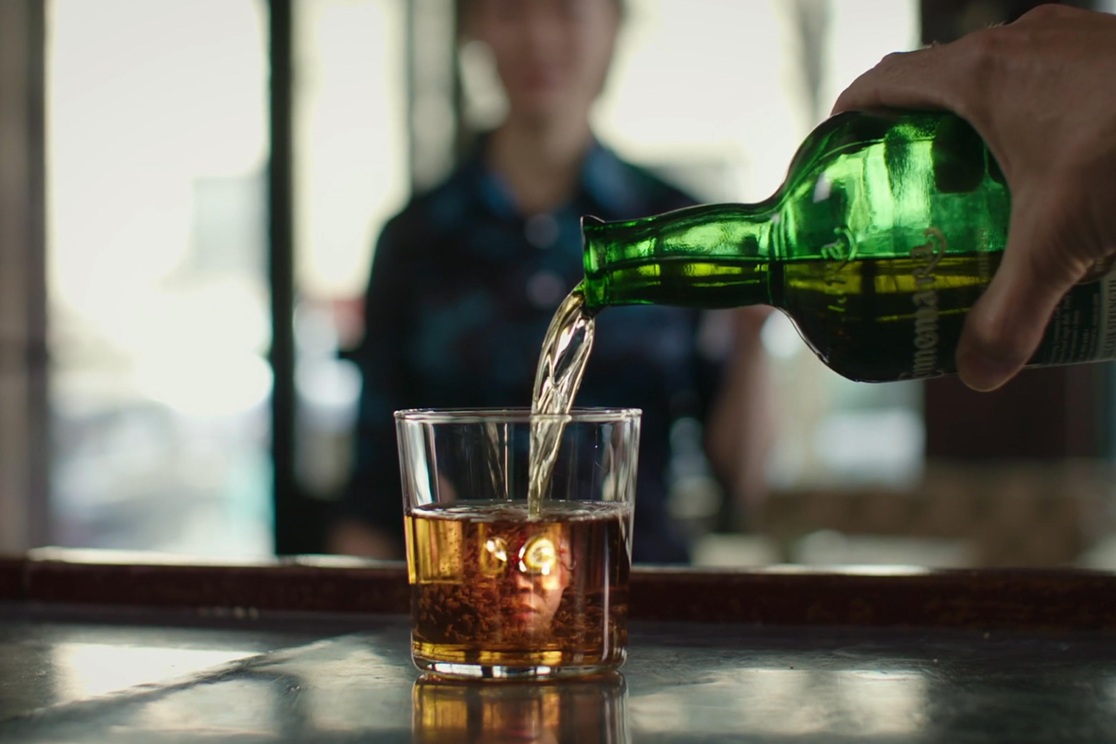 A hand pouring alcohol from a green bottle into a glass with a reflection of a face. Blurry background with a person standing.