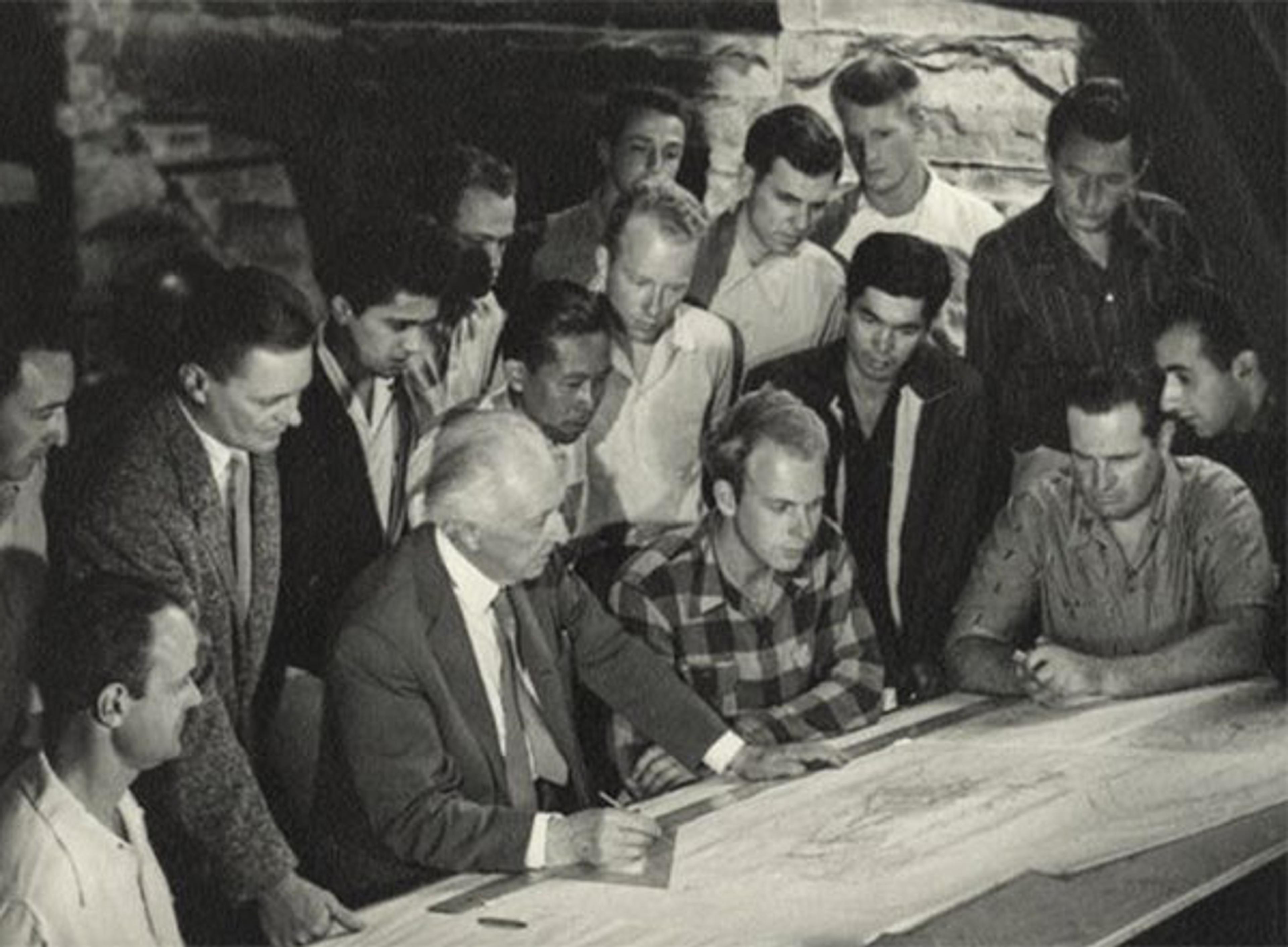 Historic black-and-white photo of a group of men gathering around a table, examining architectural blueprints.