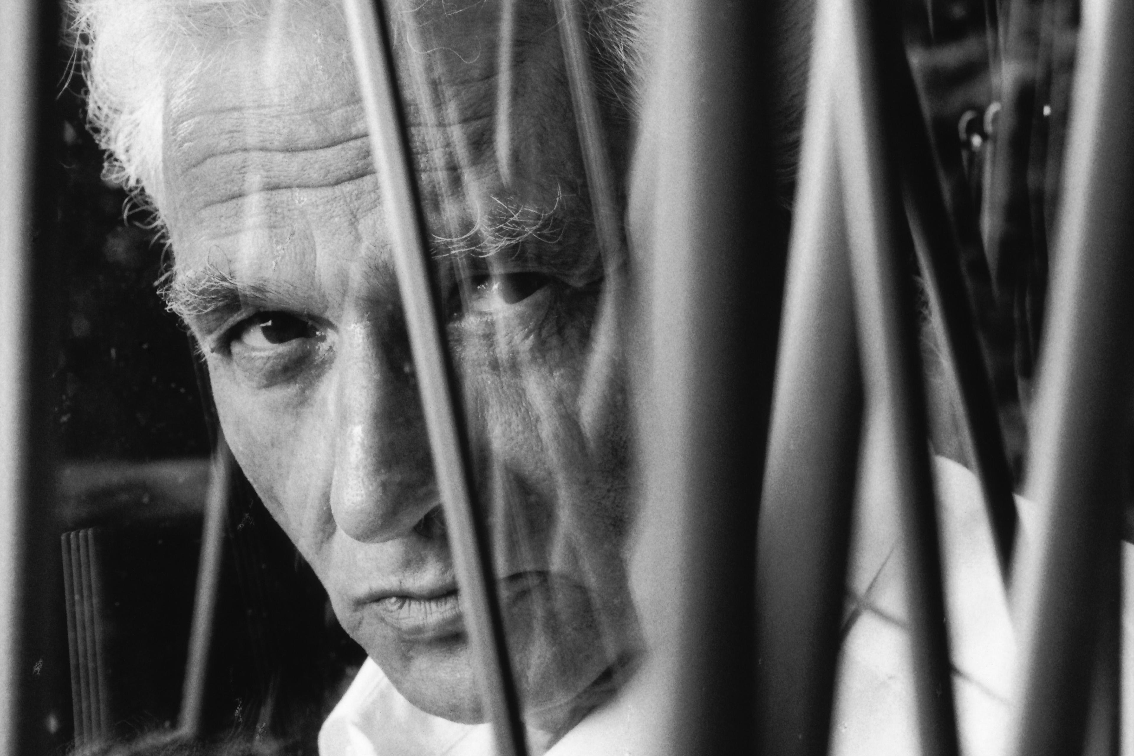 Black and white photo of a man with white hair seen through abstract lines looking intently at the camera.