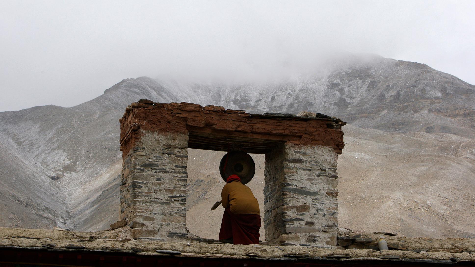 A person kneeling in front of a gong in an ancient stone structure, with a misty mountain landscape in the background.