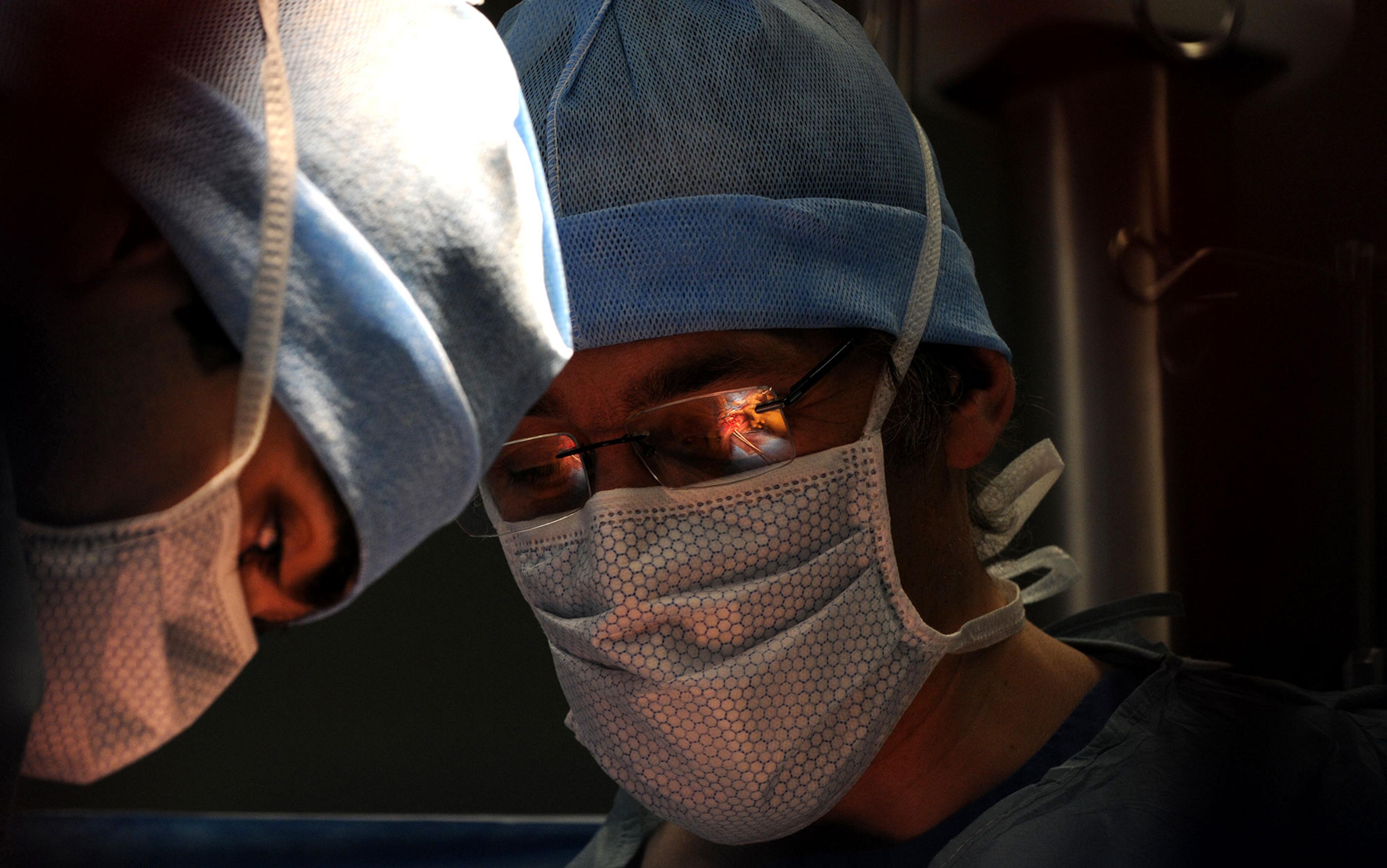 Photo of two surgeons in blue scrubs and masks focused on a procedure, with one surgeon’s glasses reflecting the surgery.